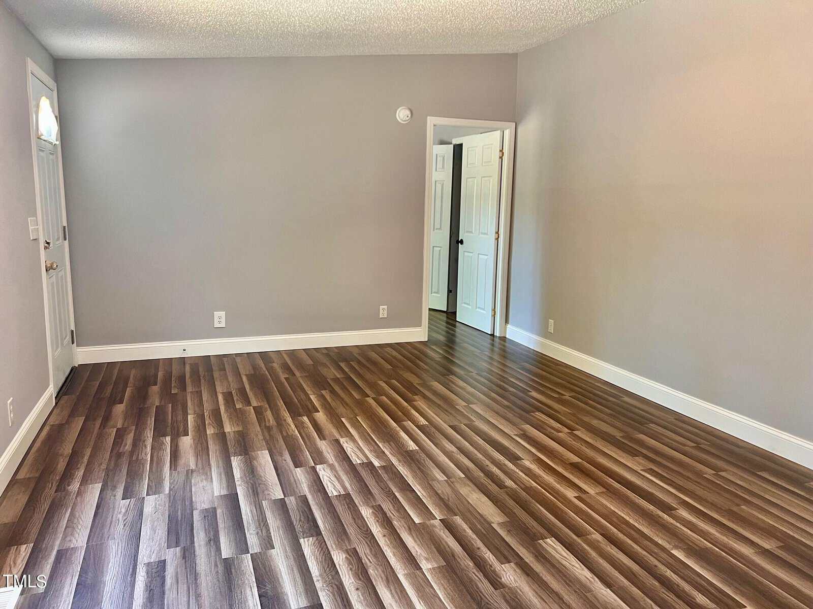 An empty room with light gray walls and dark wood flooring. A door is partially open, revealing another area. A small window near the door allows some natural light into the space.