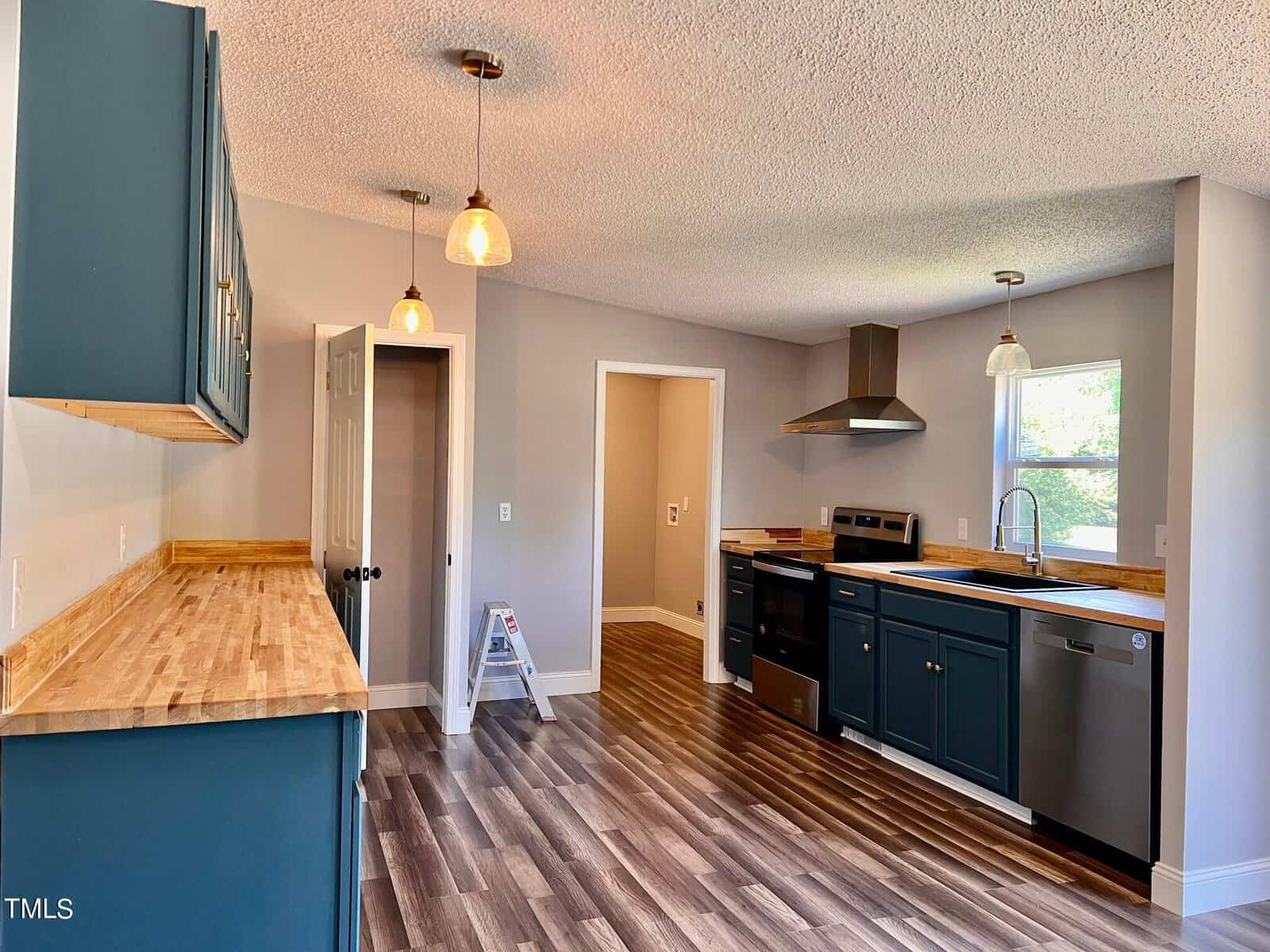 A modern kitchen with dark teal cabinets, light wood countertops, and stainless steel appliances. Pendant lights hang from the ceiling, and a step ladder is by the open doorway. The floor is wood, and natural light comes through a window.