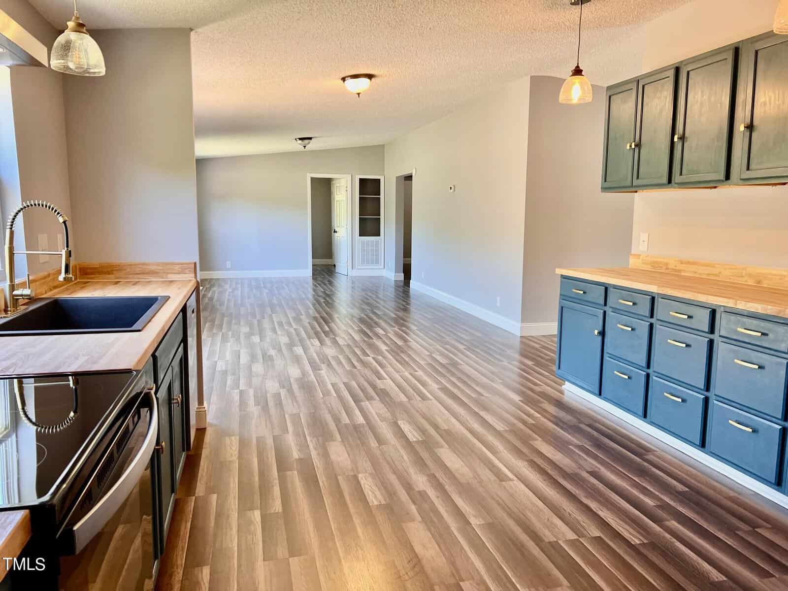 Spacious kitchen with wooden flooring, featuring a sink and black stove on the left, and blue cabinets with wooden countertops on the right. The room extends into a bright, open living area. Ceiling lights add warmth to the modern design.