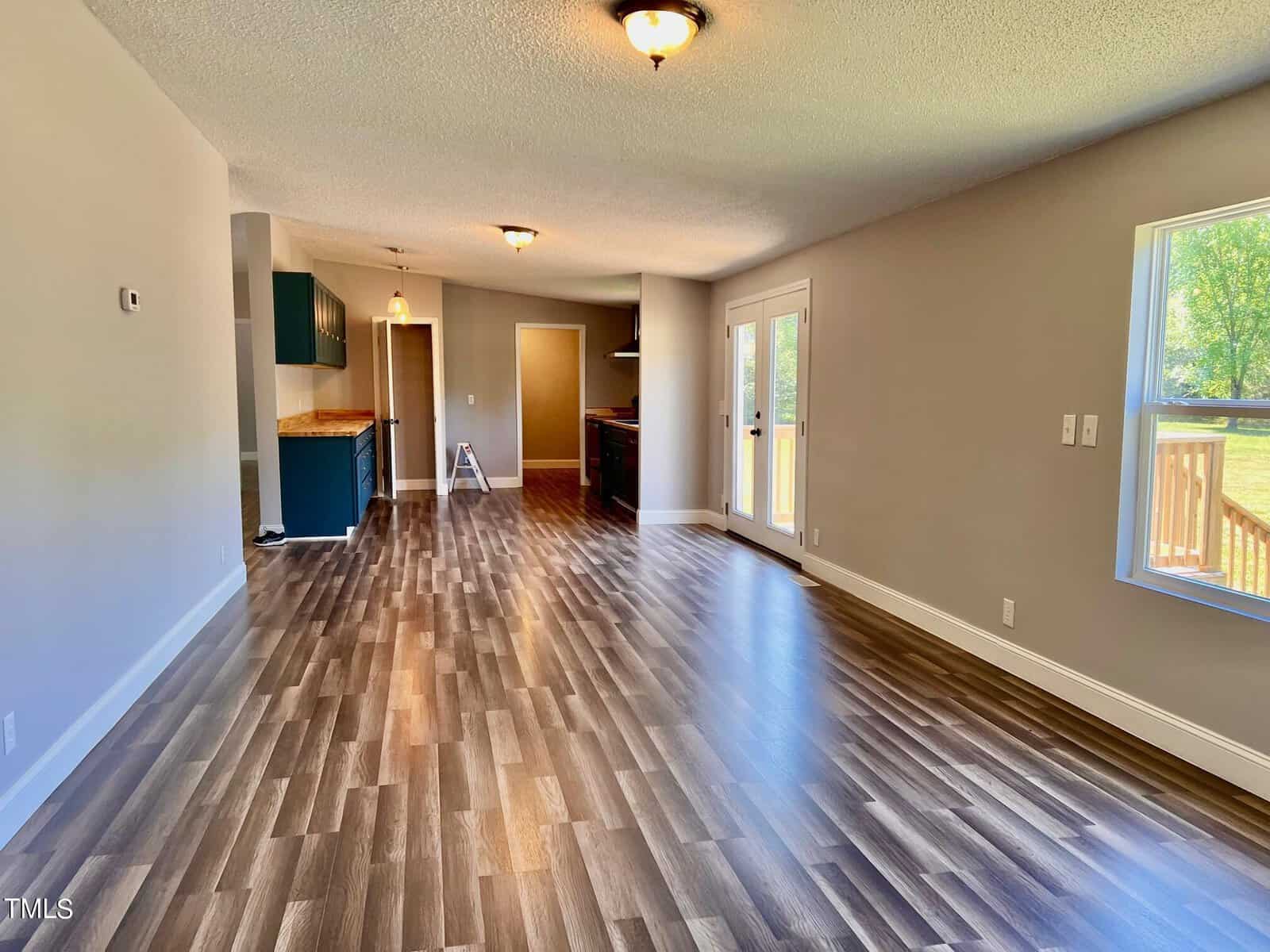 A spacious, open-concept living area with wood-style flooring and neutral walls. The room has large windows and glass doors leading outside, providing natural light. A kitchen with dark cabinets is visible in the background.