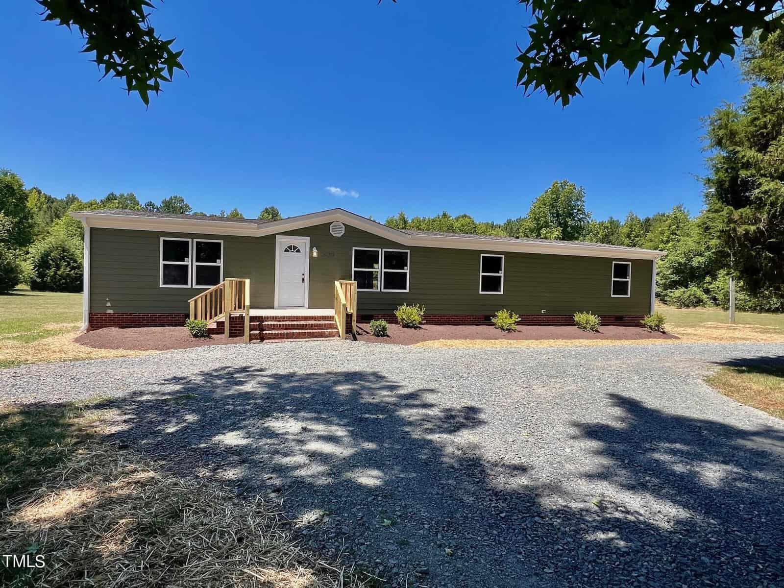 Located at 829 Tungsten Mine Road, this single-story green house with a white door and small front steps is surrounded by a well-maintained lawn and shrubs under Bullock's clear blue sky. A gravel driveway guides you to the home, framed by trees in the background.