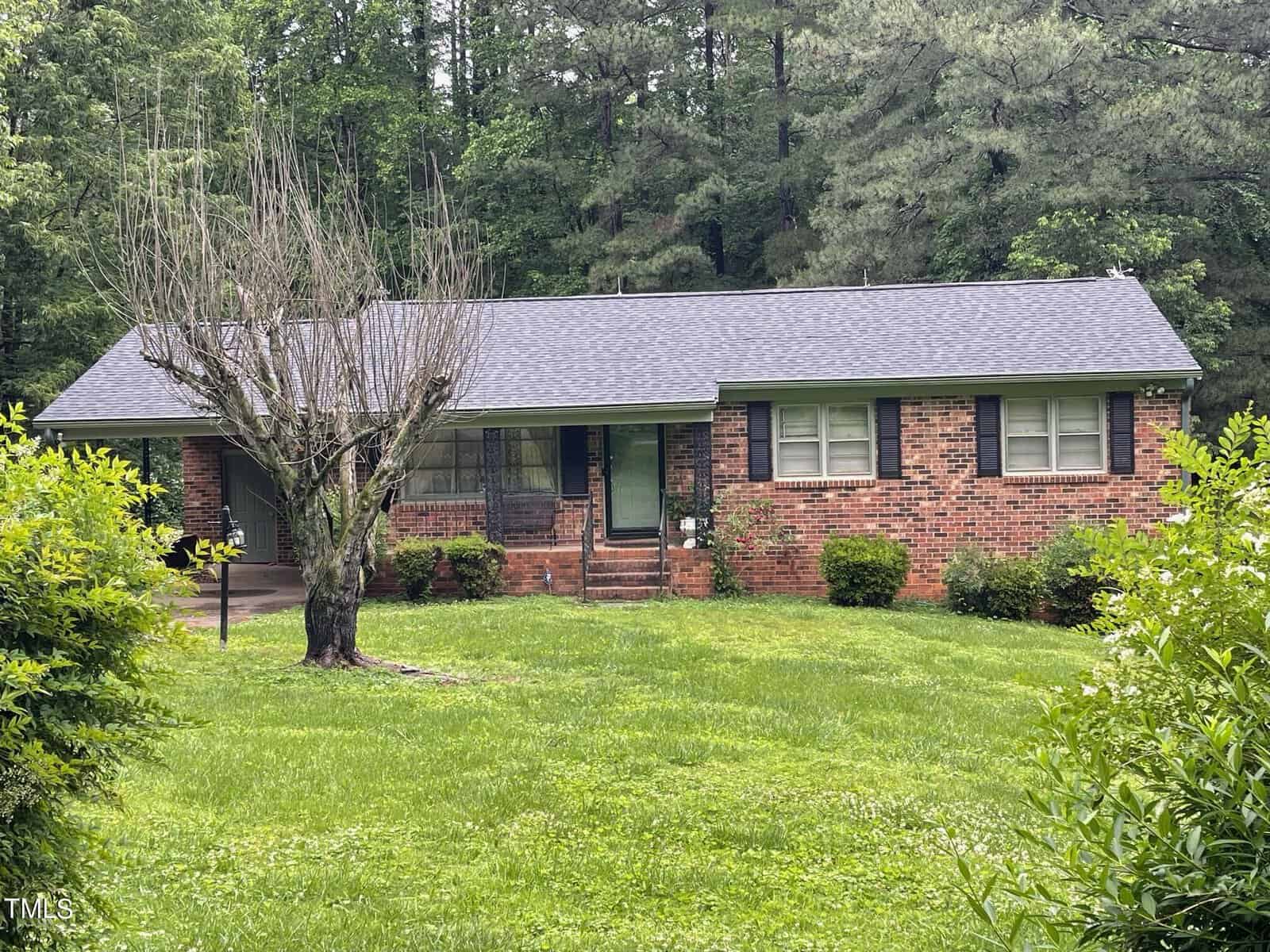 A single-story brick house with a gabled roof and a small front porch. The front yard is grassy with a few shrubs and a leafless tree near the driveway. The background features a dense forest of tall green trees.