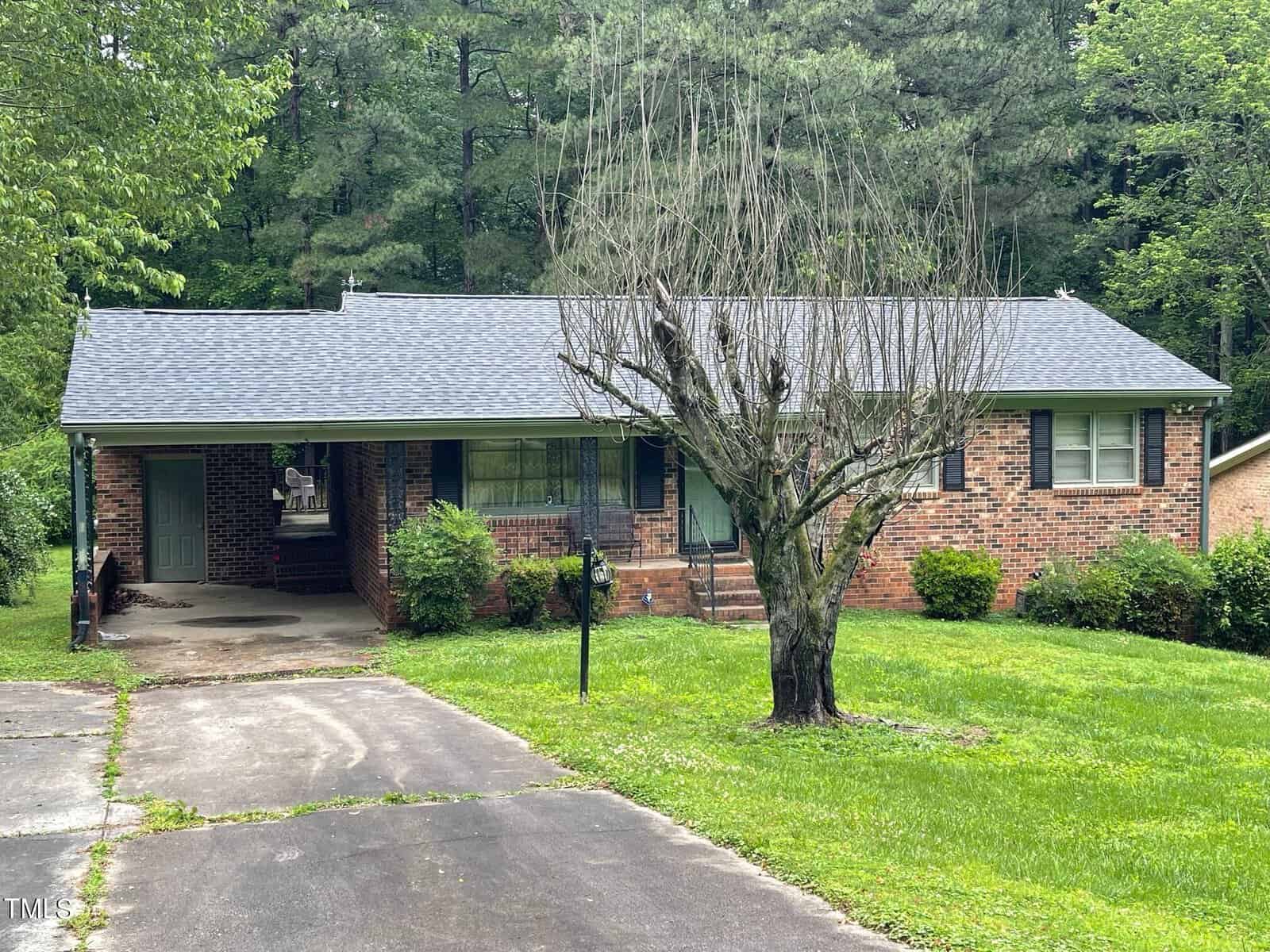 A single-story brick house with a dark gray shingle roof. The front yard features a tree with sparse branches, a well-manicured lawn, and a black mailbox. There is a driveway to the left leading to a carport, and the house is surrounded by dense, green foliage.