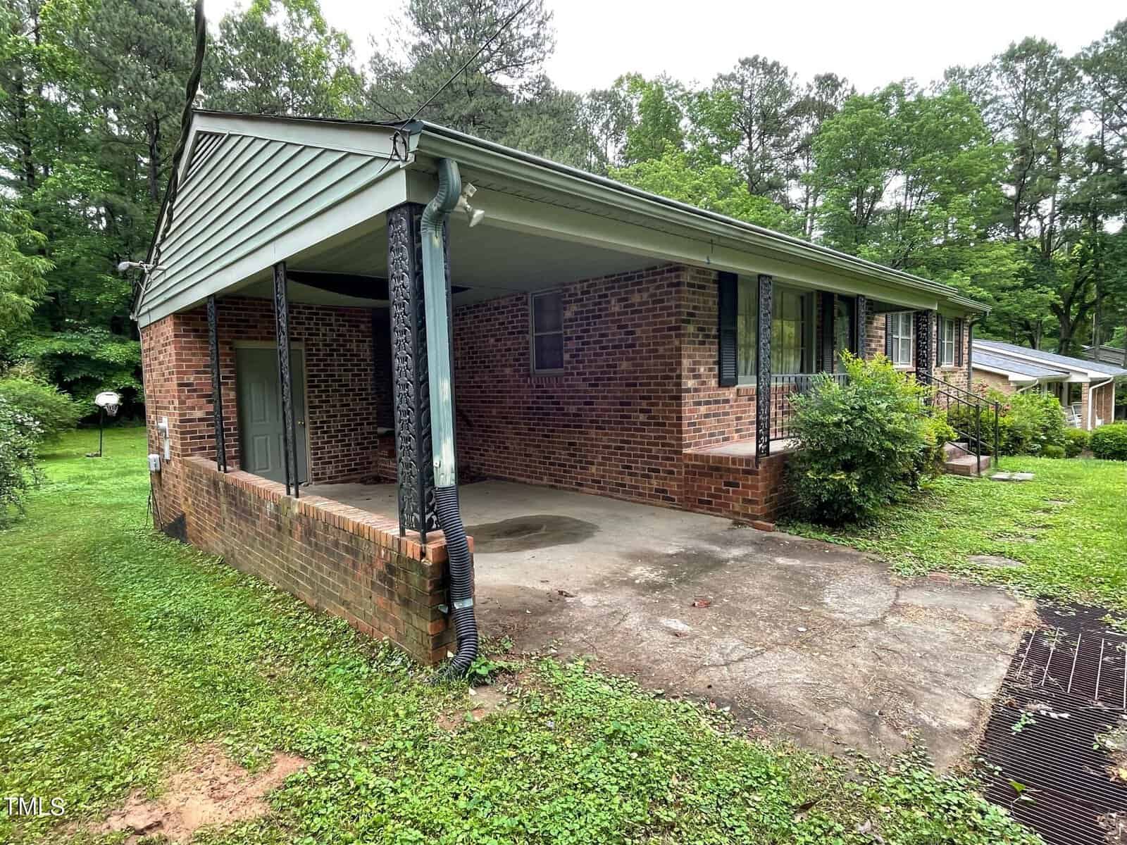 A brick house with a small front porch and an attached carport on a grassy lot. The carport has a concrete floor and is supported by black metal posts. The house is surrounded by tall trees and lush greenery, with another house partially visible in the background.