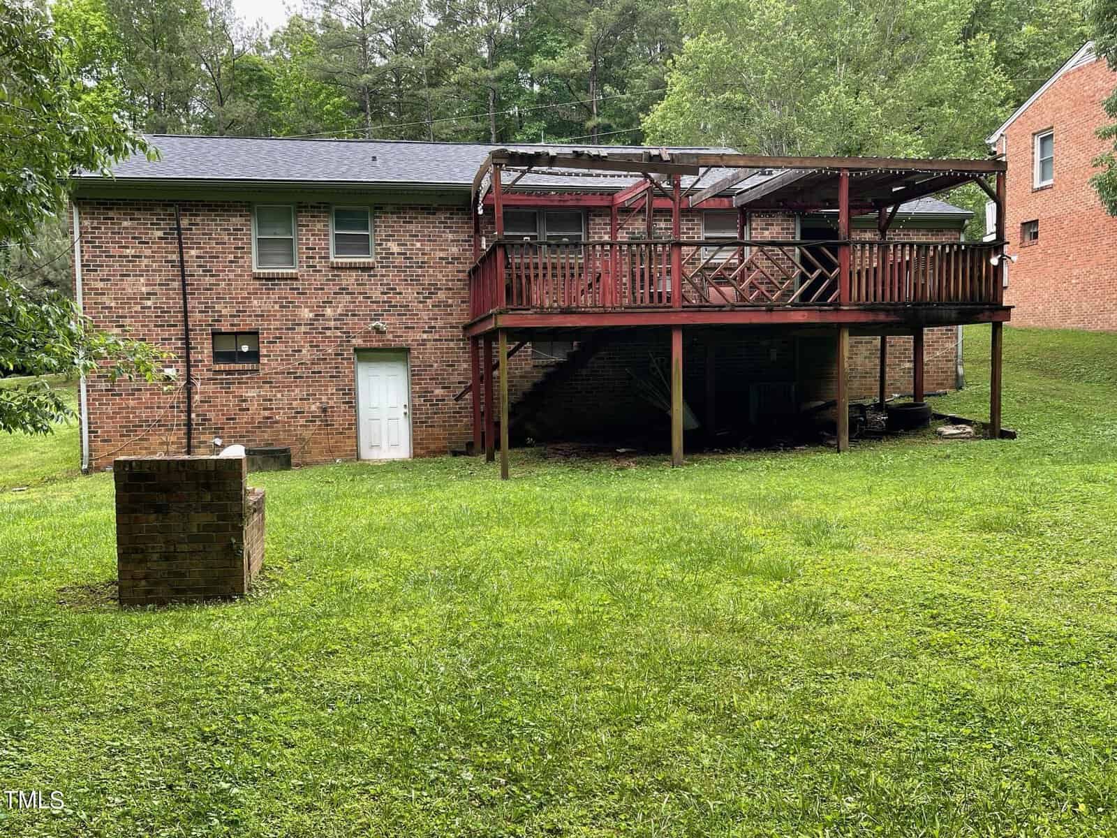 A two-story brick house with a large wooden deck at the back, elevated on tall supports. The deck is partially covered with a wooden pergola. The backyard is grassy and bordered by trees. A standalone brick structure is in the foreground on the left side.