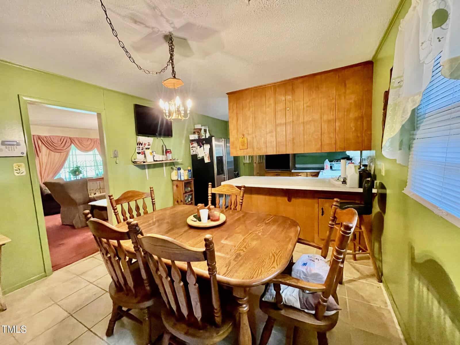 An inviting kitchen with green-painted walls features a wooden dining table and six matching chairs. A chandelier hangs above the table, which is adorned with candles. The kitchen counters and cabinets are wooden. A doorway leads to a cozy living room with red carpeting.