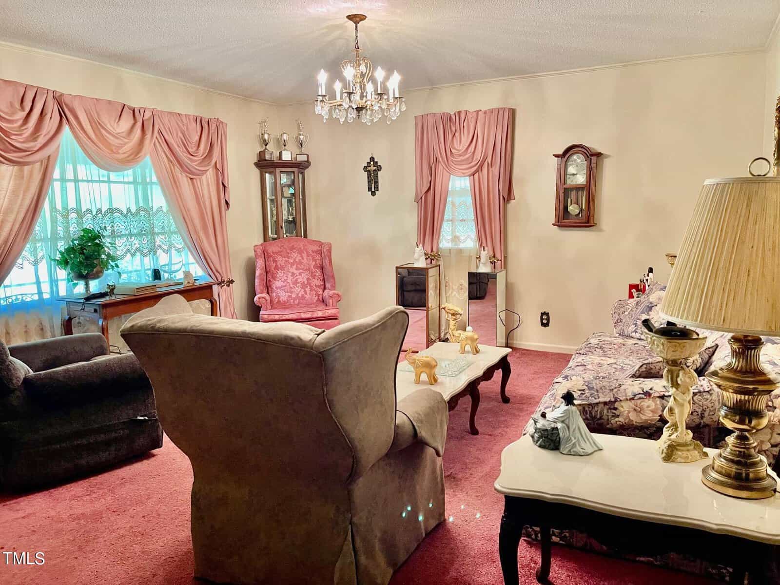 A living room with vintage decor featuring a large brown recliner, a pink armchair, a floral-patterned couch, and a dark chair. There are pink curtains, a chandelier, decorative items on tables, and wall decorations, all set on a pink carpet.
