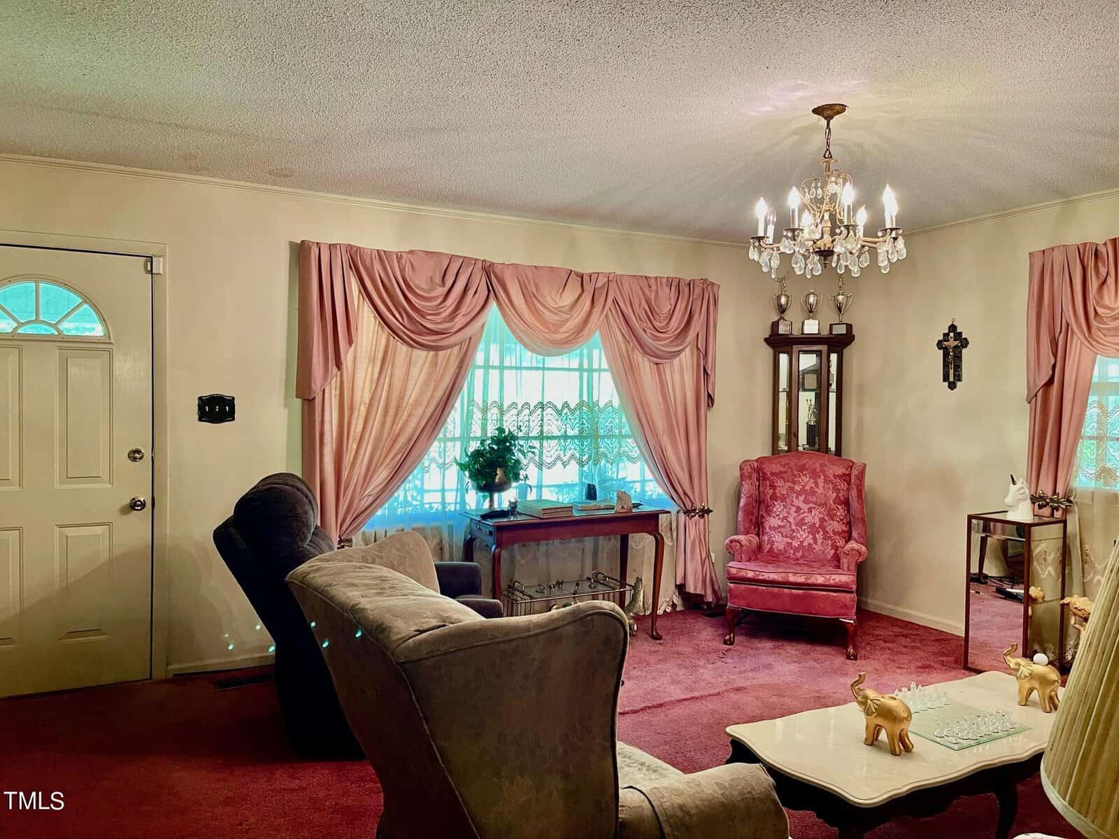 A cozy living room with a vintage feel. Pink drapes adorn the windows, and a chandelier hangs from the ceiling. There are armchairs, a small table, a pink upholstered chair, and a mirror on the wall. The door is decorated with an oval window.