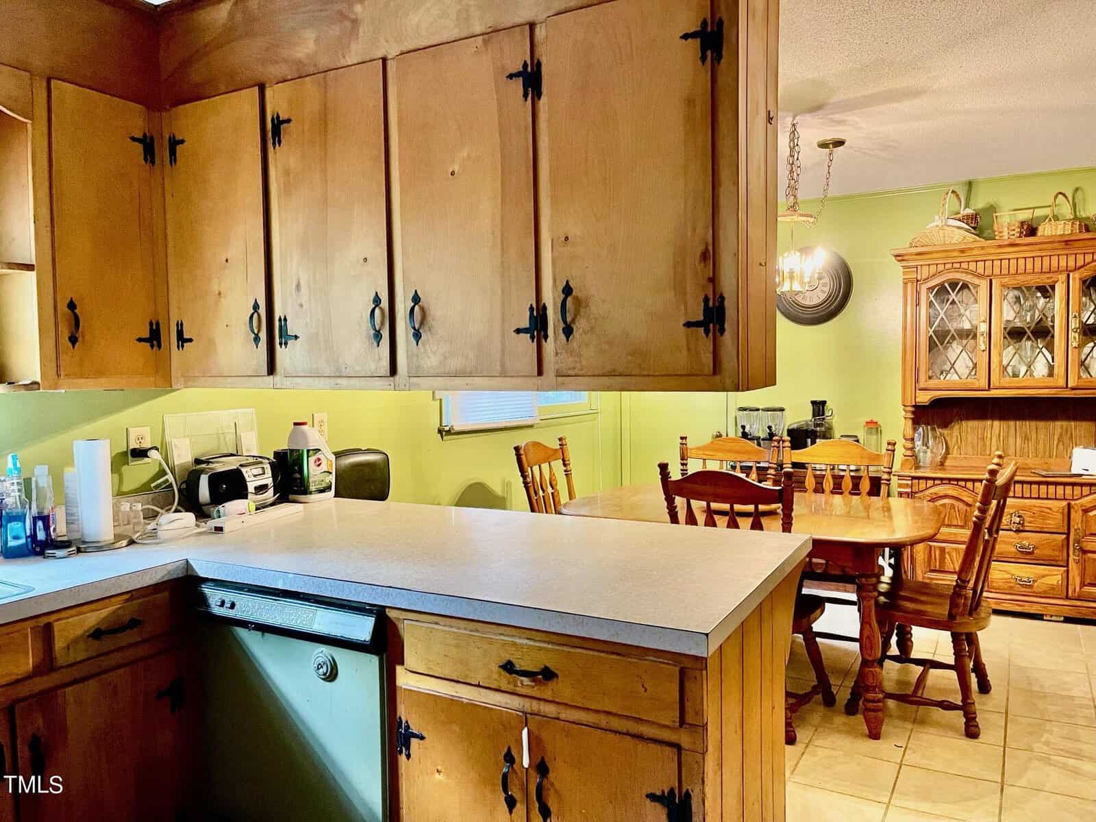 A cozy kitchen with wooden cabinets and a light countertop. A green wall with a window is visible, and the floor is tiled. Appliances and kitchen items are on the counter. In the adjacent dining area, a wooden table and chairs, along with a hutch, are seen.
