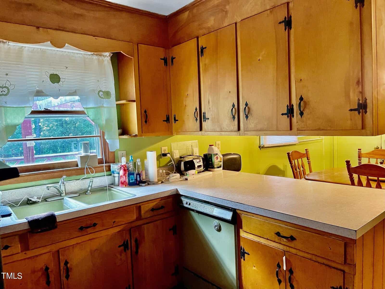 A kitchen with wooden cabinets, green counter backsplash, and a double stainless steel sink under a window with curtains. Various kitchen appliances, including a toaster and blender, are on the countertops. A dining table with wooden chairs is visible in the background.