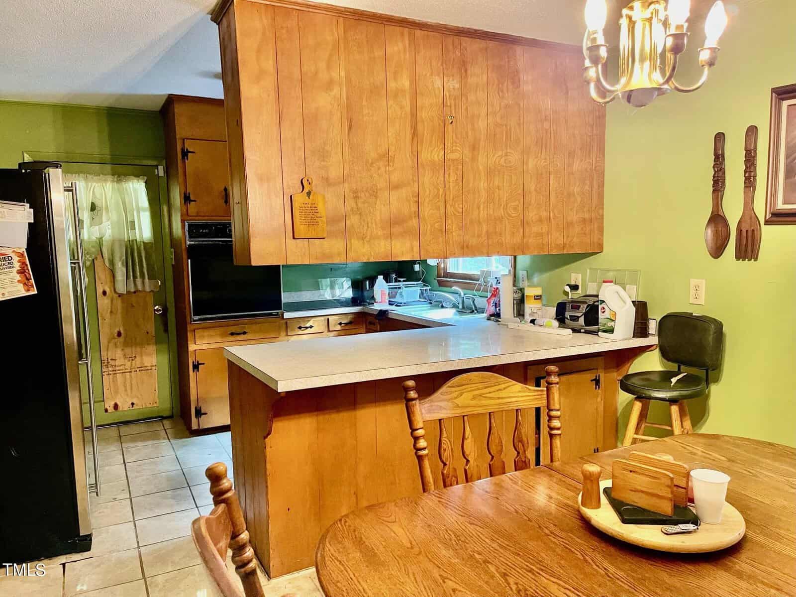 A cozy kitchen with wooden cabinets and green walls. The kitchen features a marble countertop with various appliances, including a microwave and coffee maker. Adjacent is a dining area with a wooden table and chairs, under a chandelier.