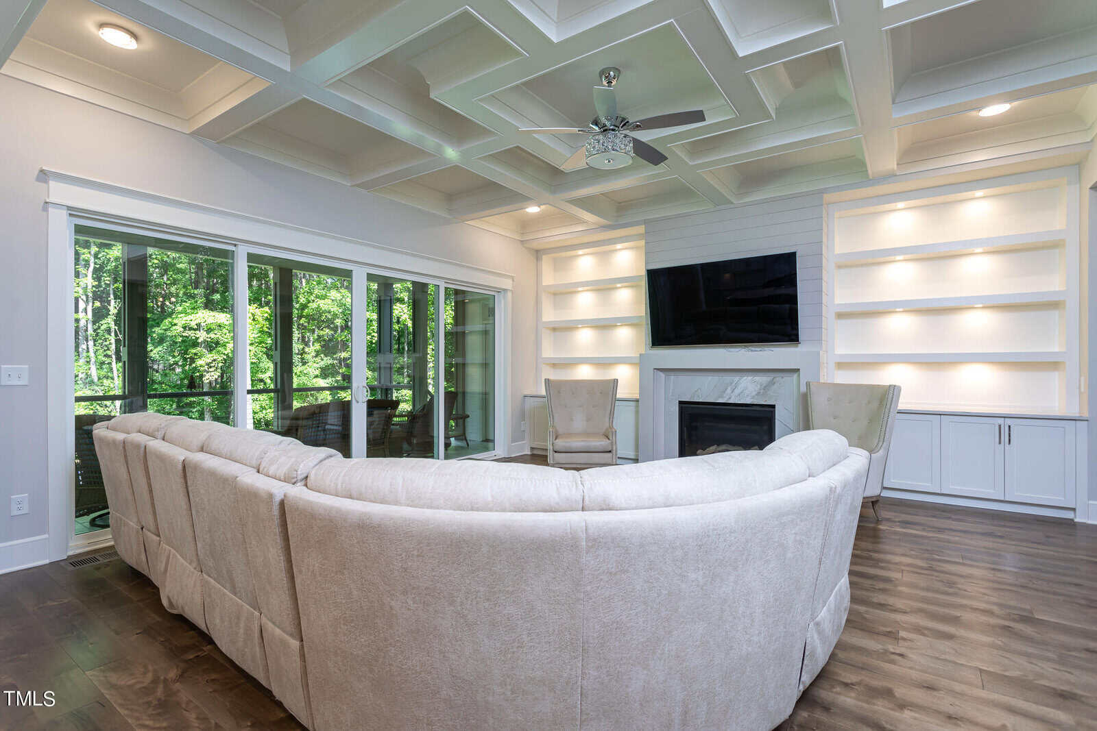 A spacious living room features a large beige sectional sofa facing a wall-mounted TV above a fireplace. Built-in shelves surround the TV. Large windows and a sliding glass door offer views of greenery outside. The coffered ceiling adds elegance.