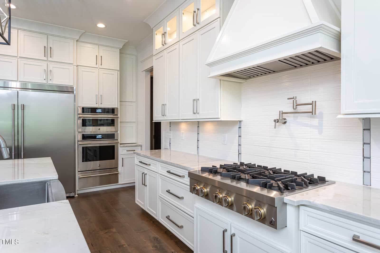 A modern kitchen with white cabinets, a stainless steel double oven, a large refrigerator, and a gas stove with a range hood. Marble countertops and a pot filler above the stove complete the sleek design.