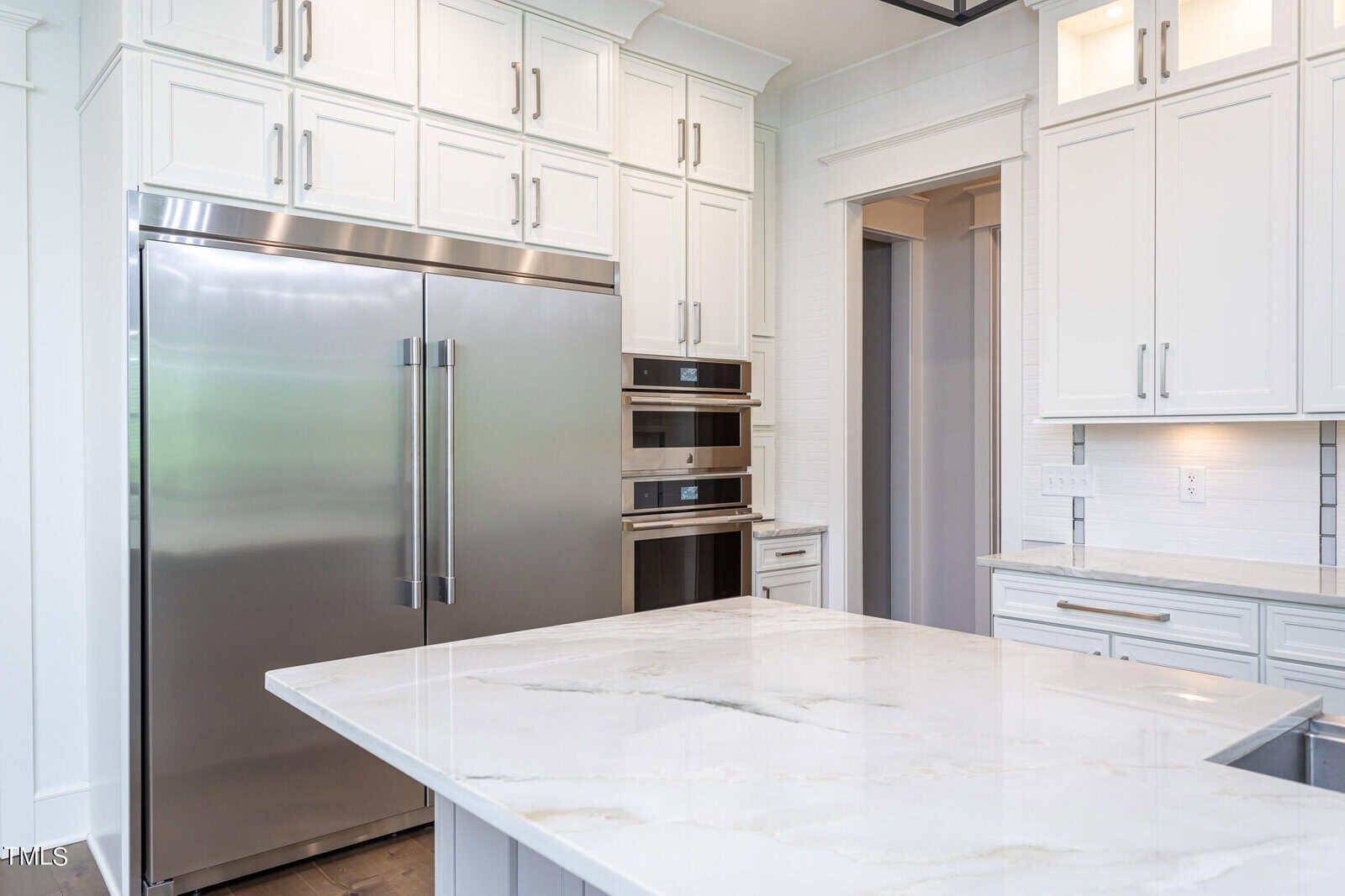 A modern kitchen featuring a large stainless steel refrigerator, double wall ovens, white cabinetry, and a spacious marble island countertop. The room is bright with ample natural light.