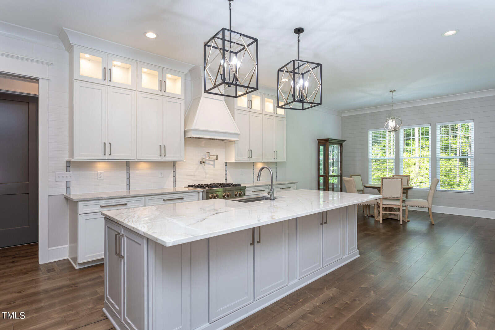 A modern kitchen with white cabinets, a large marble island, two geometric pendant lights, and a sink. Hardwood floors extend to a dining area with a table and chairs near large windows. A glass cabinet stands in the corner.