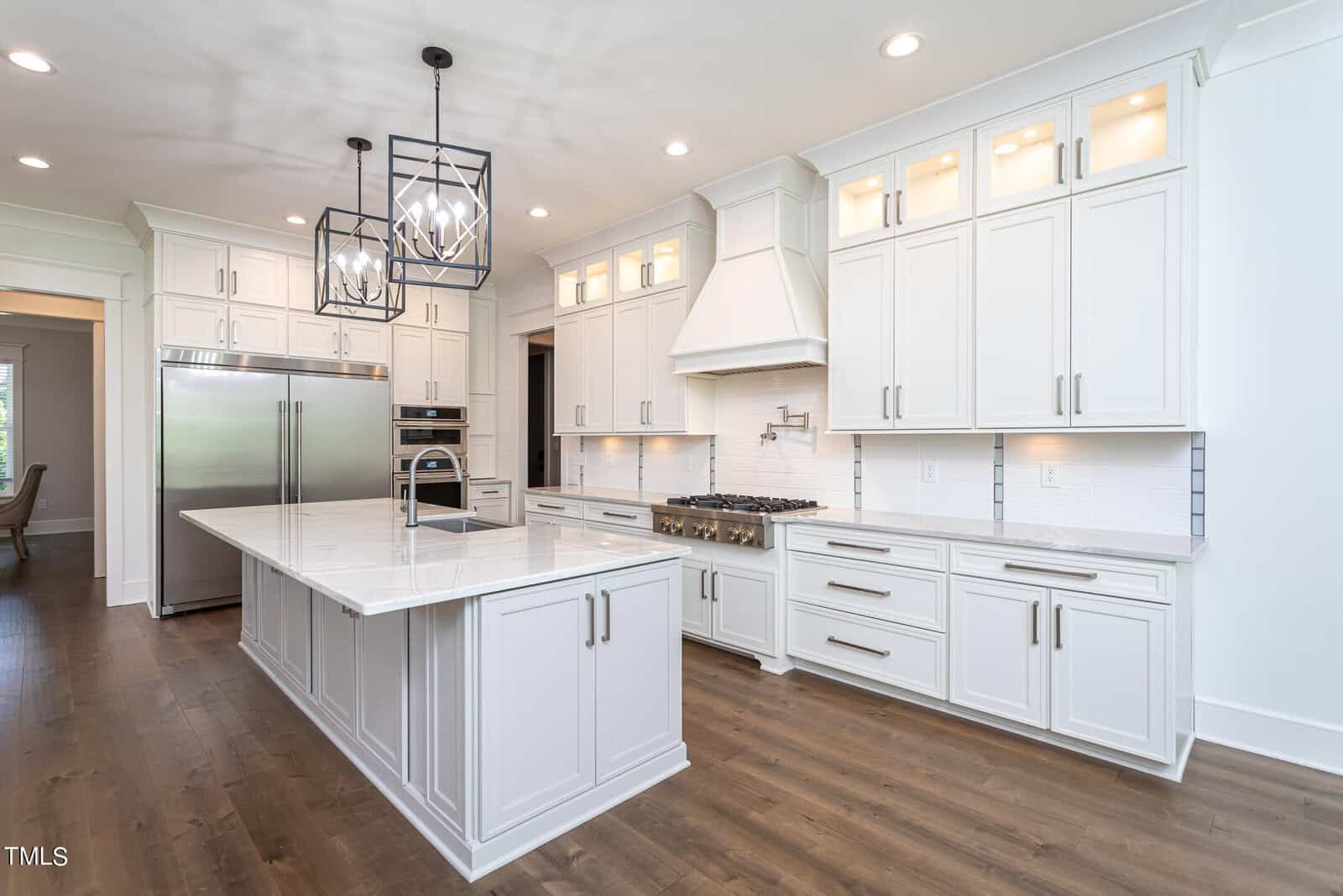 Modern kitchen with white cabinets, island with marble countertop and sink, stainless steel appliances, and dark wood flooring. Pendant lights hang above the island. The room is spacious and bright.