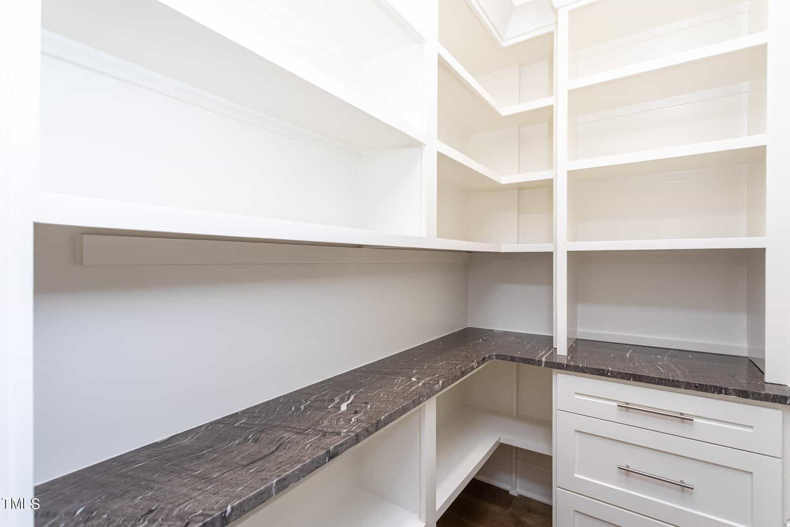 A bright and spacious corner pantry features multiple white shelves, a dark stone countertop, and white cabinetry with metal handles. The shelves and countertop create ample storage space against the light-colored walls.