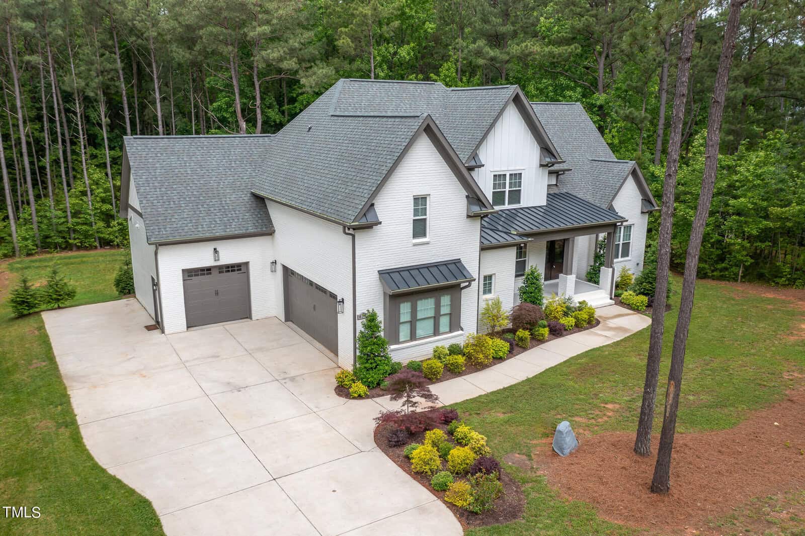 The two-story white house at 675 Willard Drive in Creedmoor boasts a gray roof and is surrounded by trees. With black window accents, it offers a spacious driveway to a two-car garage, complemented by well-maintained landscaping featuring green shrubs and a small stone garden.