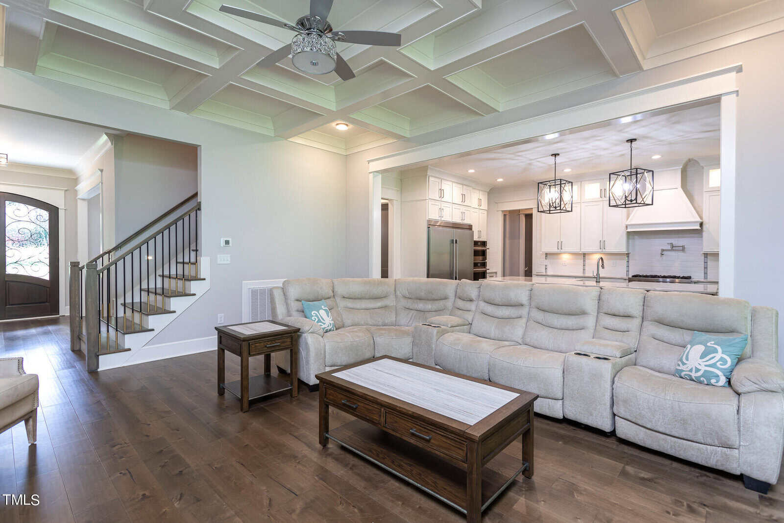 A spacious, modern living room with a coffered ceiling and wood flooring. It features a large beige sectional sofa, wooden coffee and side tables, and an open view of a white kitchen with pendant lights. A staircase is visible on the left.