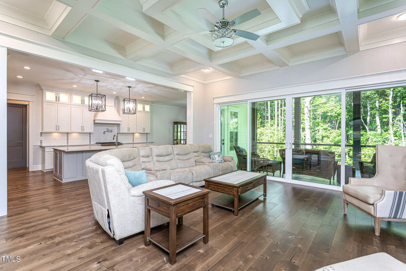 Spacious living room with coffered ceiling, light-colored sofas, and a wooden coffee table on polished wood floors. Large glass doors open to a balcony with forest views. Adjacent to an open kitchen with white cabinetry and hanging lights.
