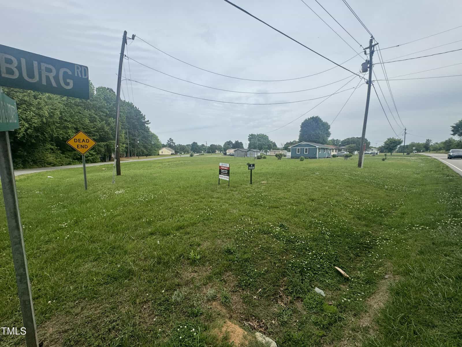 A grassy vacant lot is pictured, with a "Dead End" sign and power lines visible. There is a street sign in the foreground, houses in the background, and a "For Sale" sign on the property. The sky is overcast.