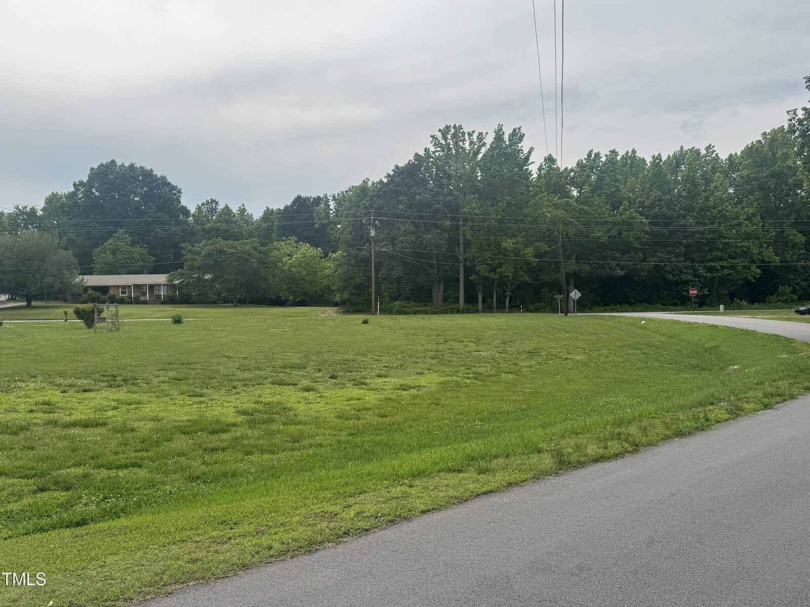 A green lawn with a few scattered trees and shrubs extends to a house in the background surrounded by more trees. In the foreground, a paved road curves to the right, with power lines and a stop sign along the roadside. The sky is cloudy and overcast.