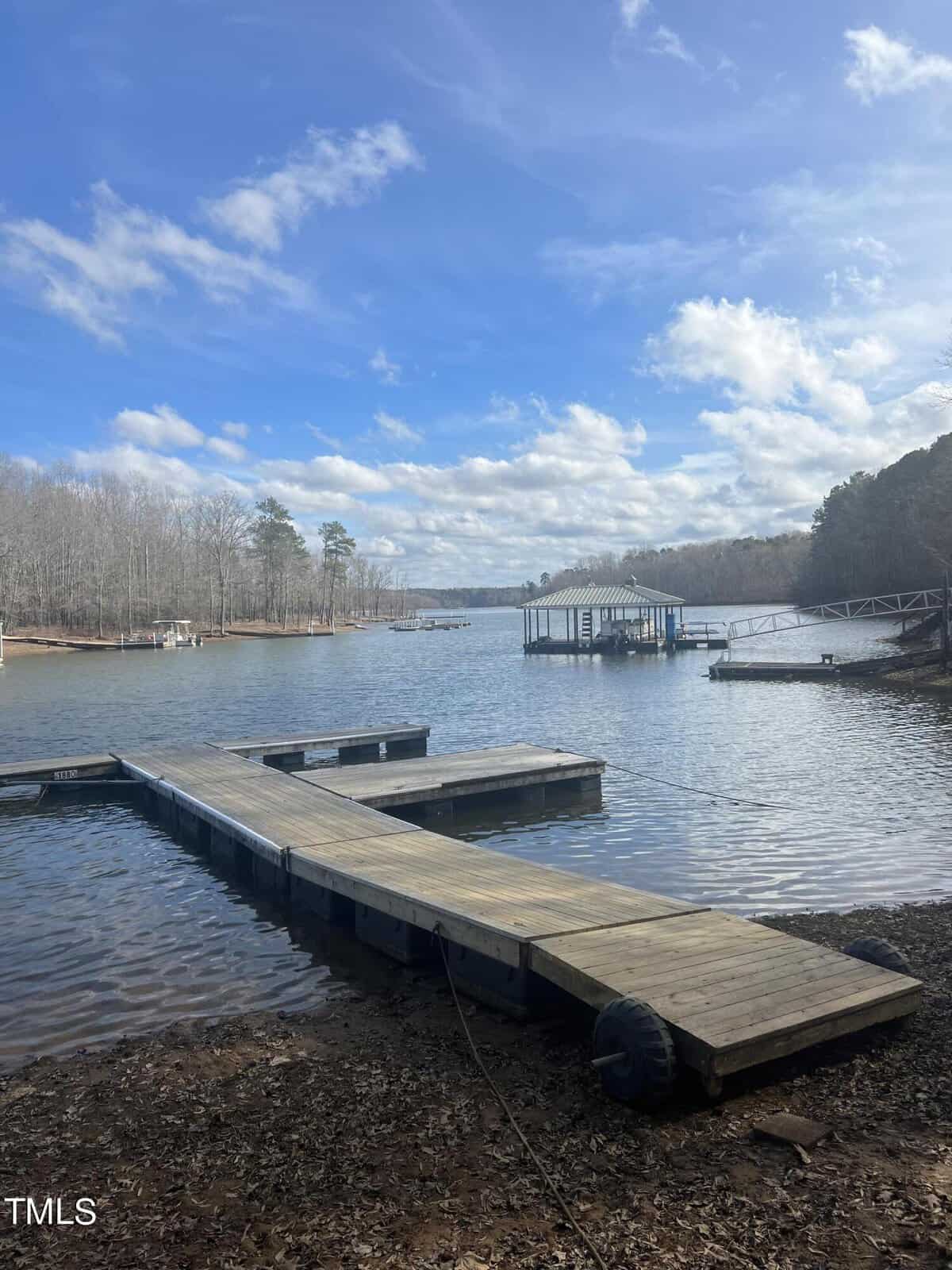 A scenic view of a calm lake with a wooden dock extending into the water under a partly cloudy sky. In the distance, there is another small dock structure with some trees along the shoreline. The area is serene and surrounded by a mix of bare and evergreen trees.