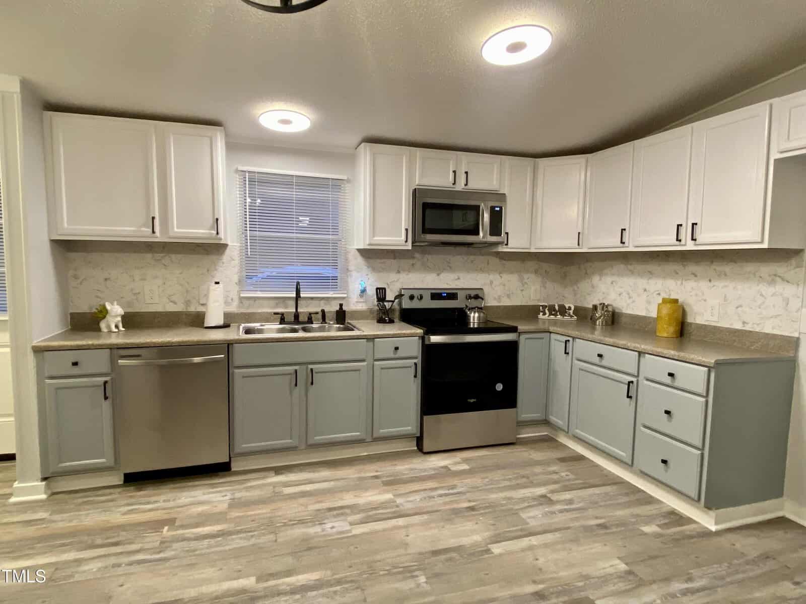 A modern kitchen with white overhead cabinets and grey lower cabinets. It features stainless steel appliances, including a dishwasher, microwave, and oven. The countertops are beige, and the backsplash has a subtle pattern. The floor is a light grey wood laminate.