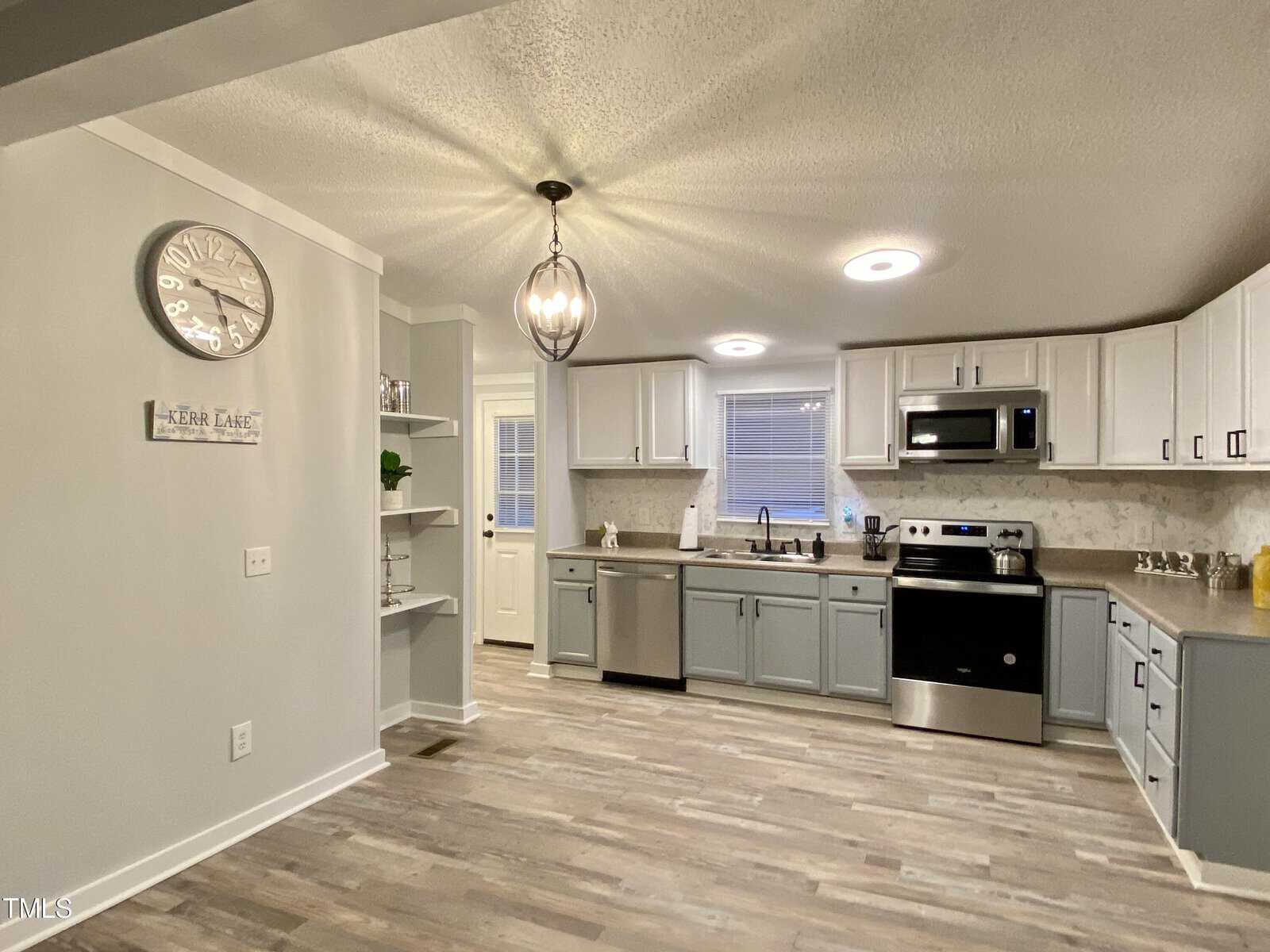 A modern kitchen with light grey walls, white upper cabinets, and grey lower cabinets. Stainless steel appliances include a stove, microwave, and sink. A decorative light fixture hangs from the ceiling. The room features wooden flooring and a wall clock.