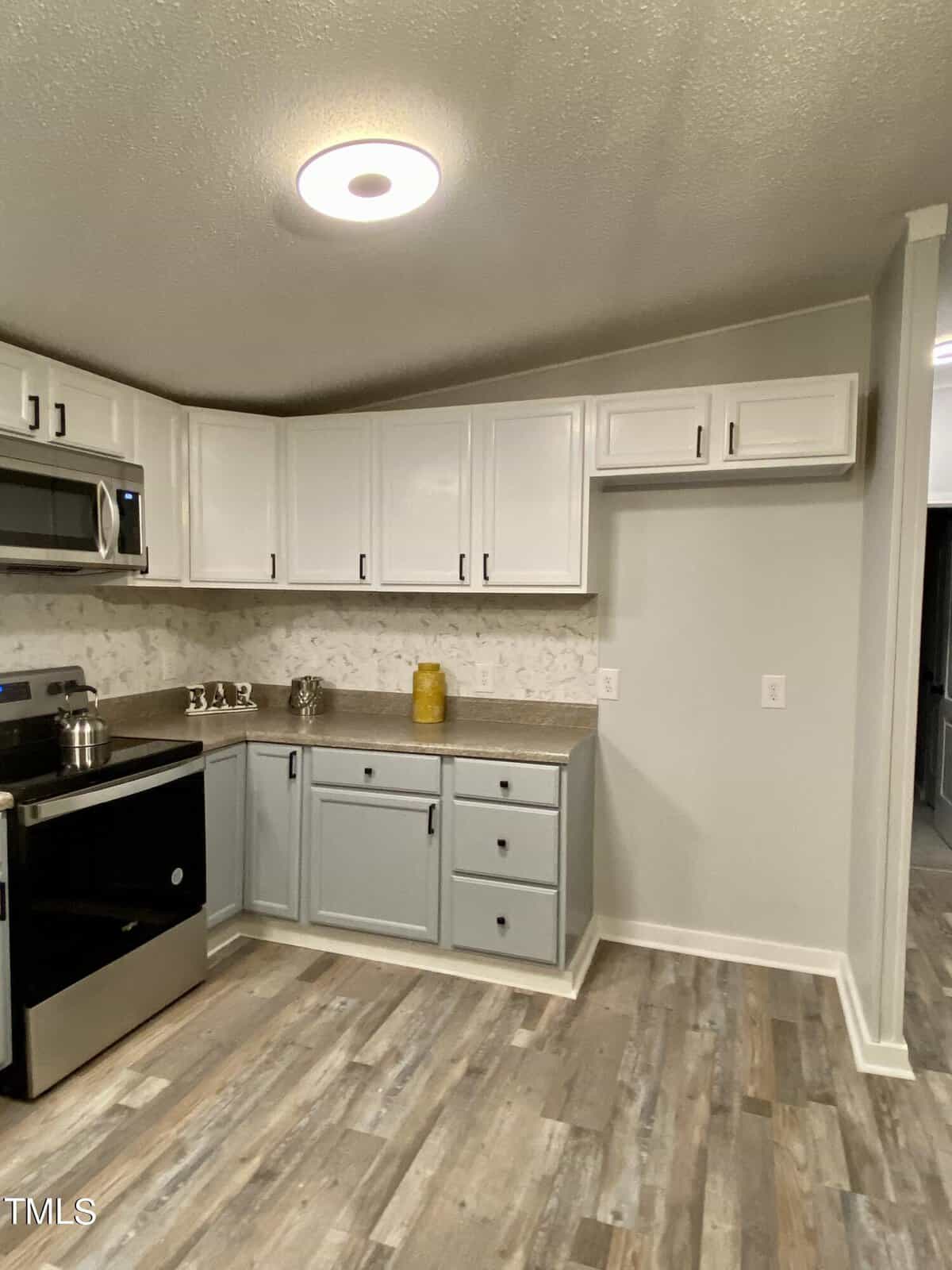 A modern kitchen with grey lower cabinets and white upper cabinets, complemented by stainless steel appliances, including a stove and microwave. The countertop features decorative items, and the floor is made of light-colored wood. The walls are painted light grey.