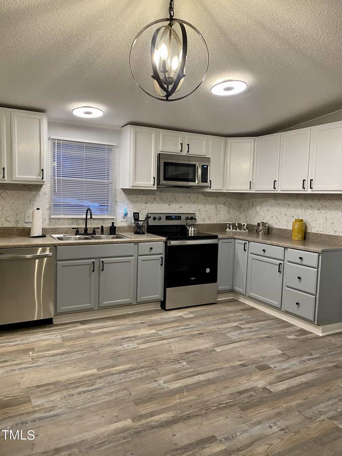 This image shows a contemporary kitchen with white upper cabinets and light gray lower cabinets. There is stainless steel appliances that include a dishwasher, stove, and microwave installed above the stove. The countertops are a granite-like material and the floor is a light wood. A modern light fixture is hanging from the ceiling.