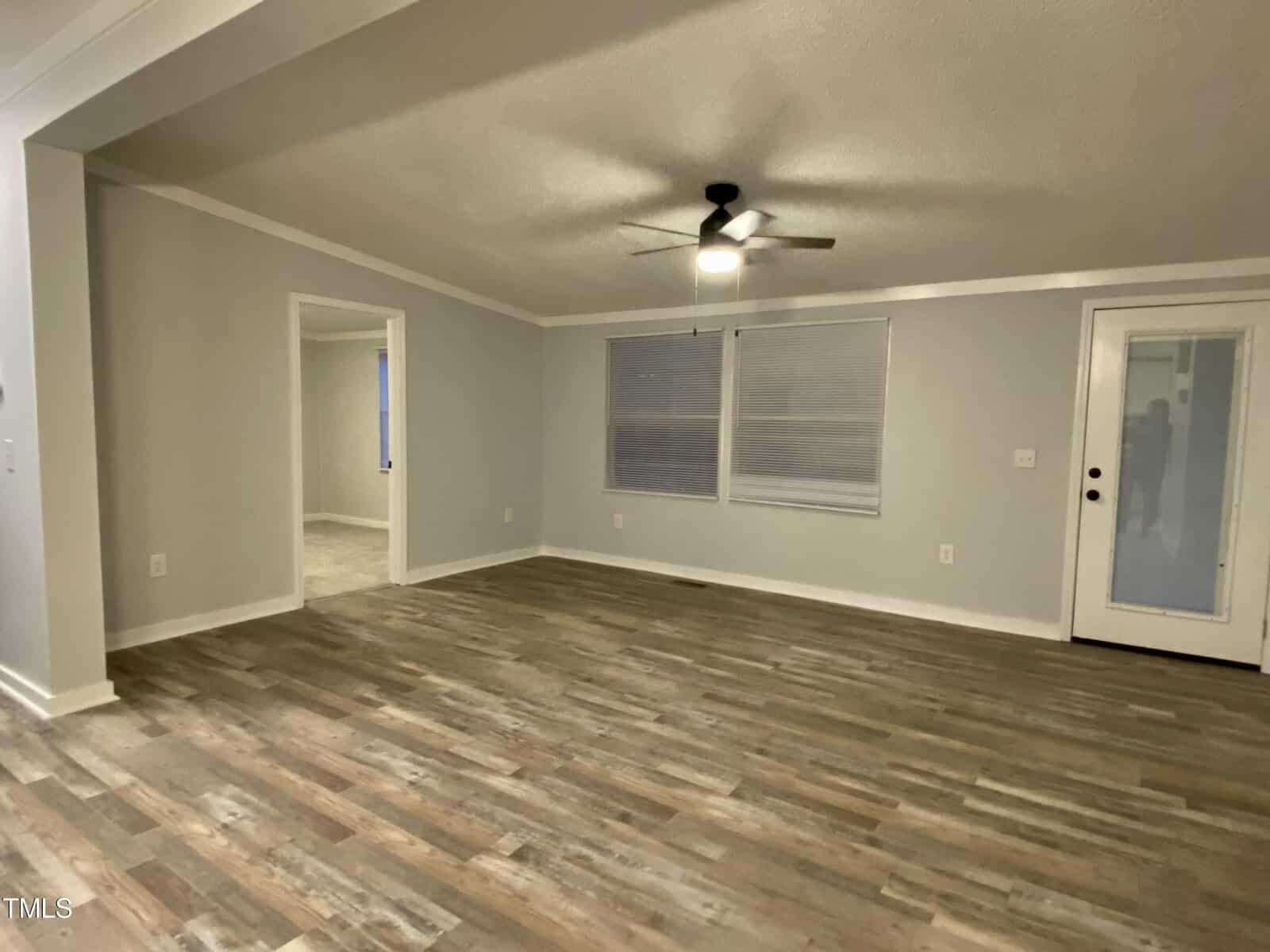 A spacious, empty living room with light gray walls and a wood-patterned vinyl floor. The room features a ceiling fan with a light, two windows with closed blinds, and a door with a glass panel leading to the outside. Another room is visible through a doorway.