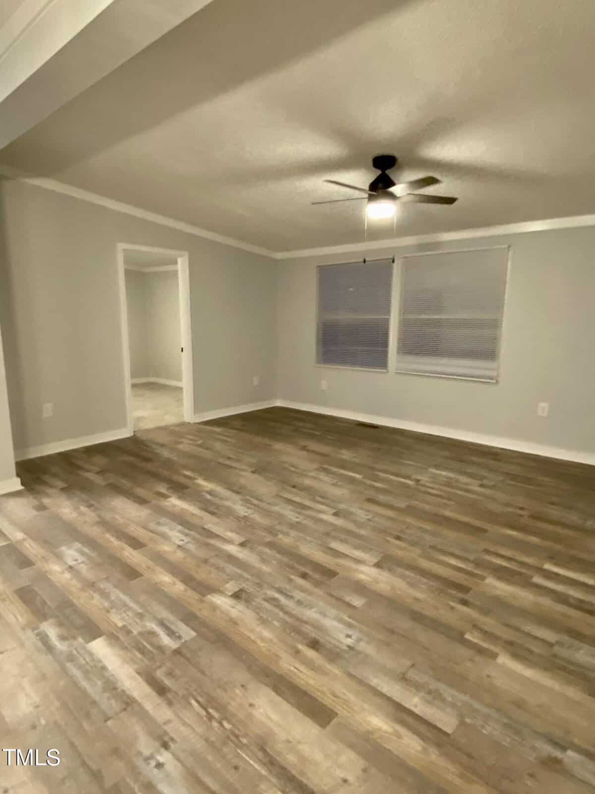 A spacious, unfurnished room with light brown hardwood flooring and light gray walls. The room features two windows with blinds, a ceiling fan with a light fixture, and an open doorway leading to an adjoining area.