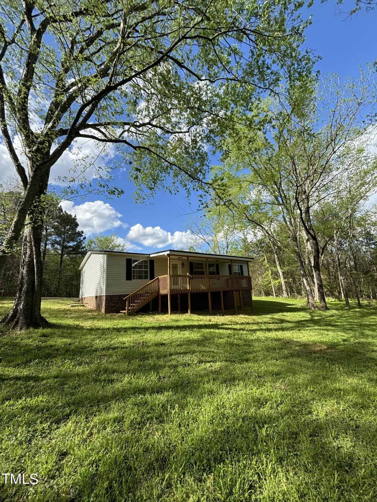 A single-story house with a wooden porch and raised brick foundation, surrounded by a grassy lawn and tall trees. The sky is blue with scattered clouds, and the sunlight is casting shadows on the ground. The house is situated in a serene, natural setting.