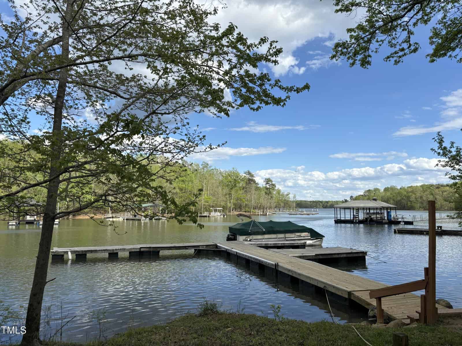 A scenic lakeside view featuring a wooden dock extending into the water with a boat covered by a green tarp. Surrounding the lake are lush, green trees under a partly cloudy sky, creating a tranquil and picturesque atmosphere.