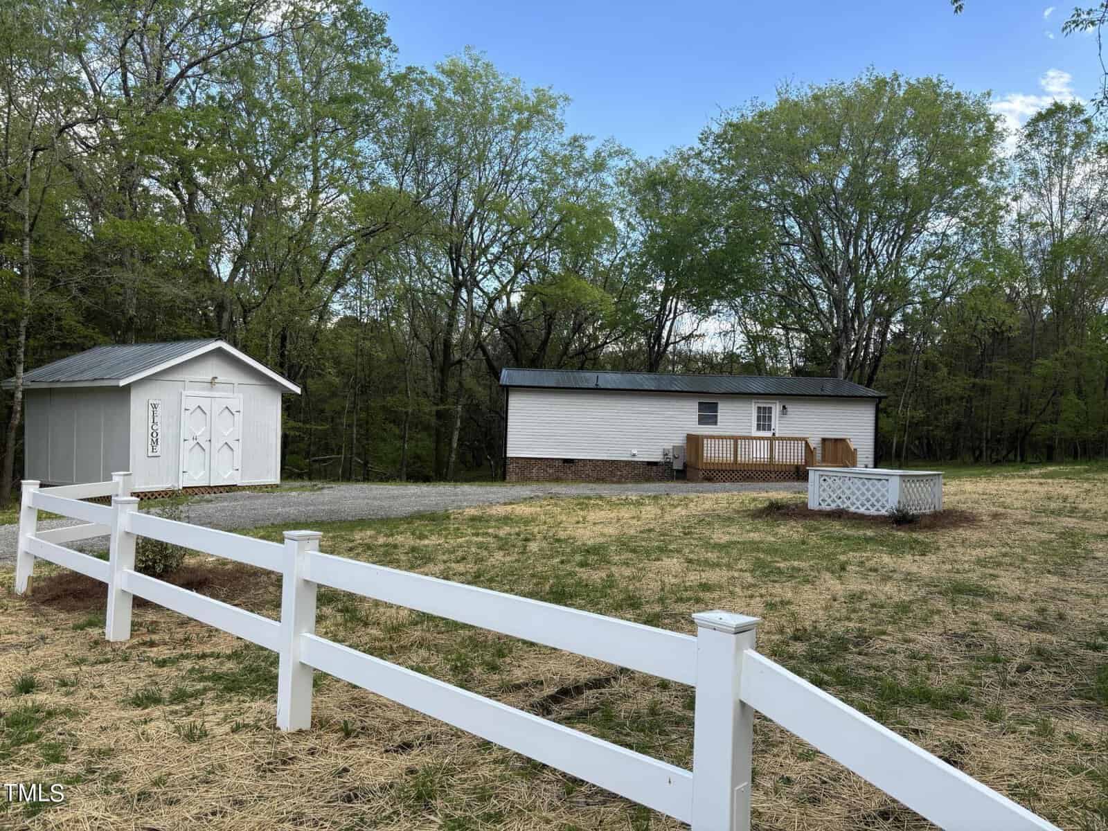 A small white house with a gray roof and brick foundation sits in a clearing surrounded by trees. Next to the house, there is an identical white shed. A white fence runs along the foreground, enclosing a grassy area. A wooden deck is attached to the house.