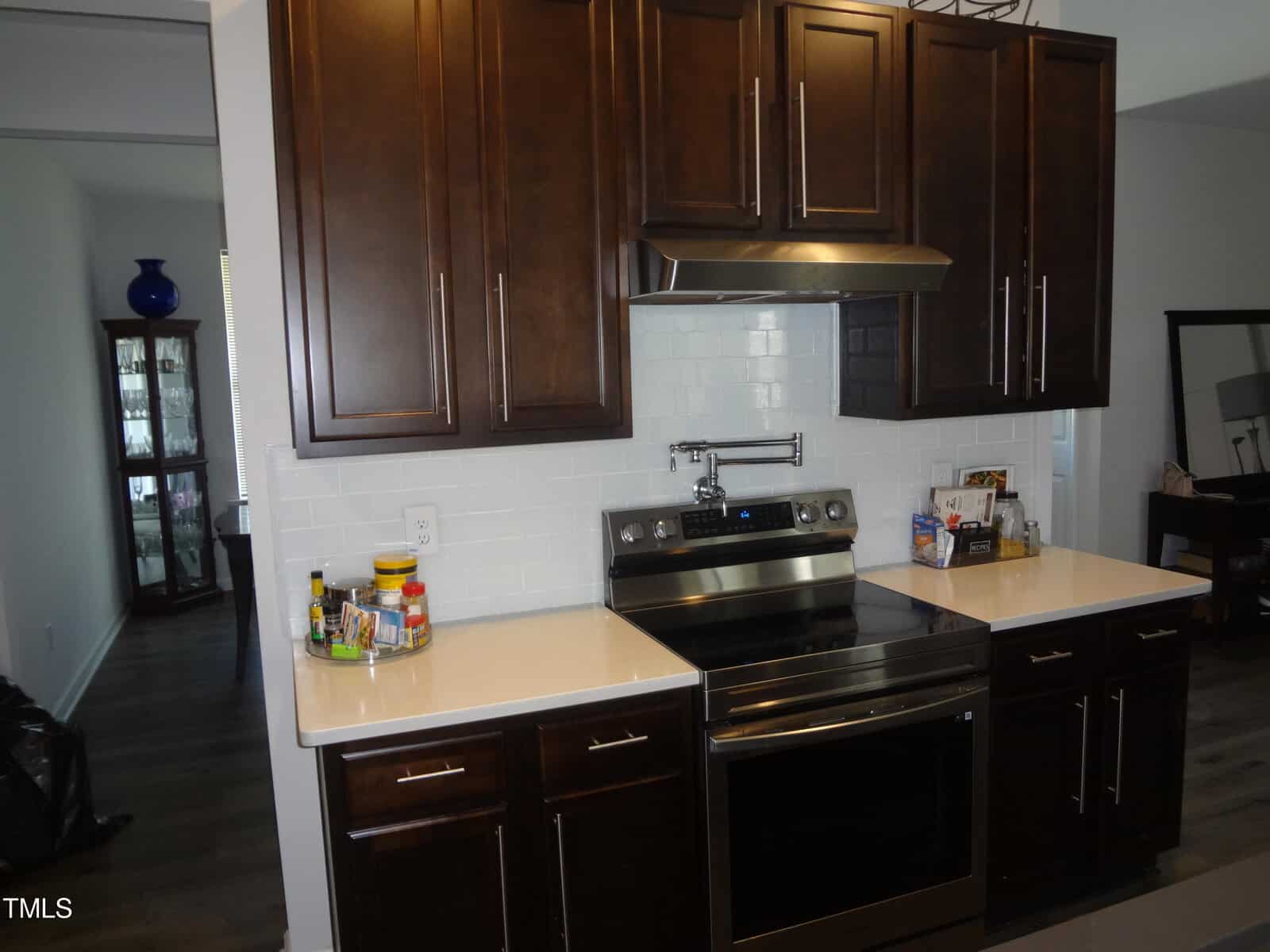 A modern kitchen at 1850 Gillburg Road, Henderson NC, features dark wood cabinets, white countertops, and a stainless steel electric range with a hood. The backsplash is white subway tile. To the left is a hallway leading to another room, and on the right, part of a dining or living area is visible.