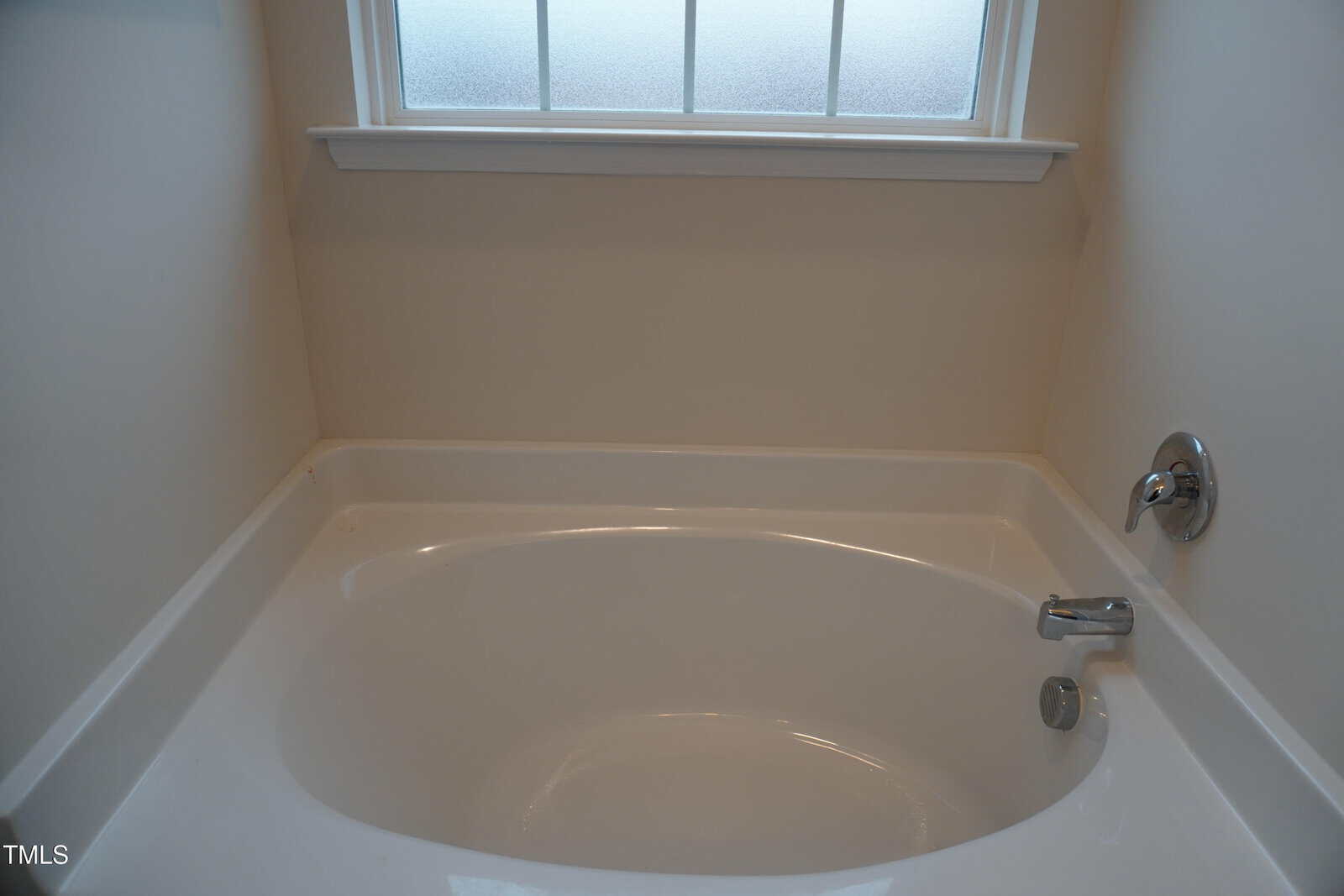 A white bathtub with silver faucet fixtures is set against cream-colored walls beneath a frosted window.