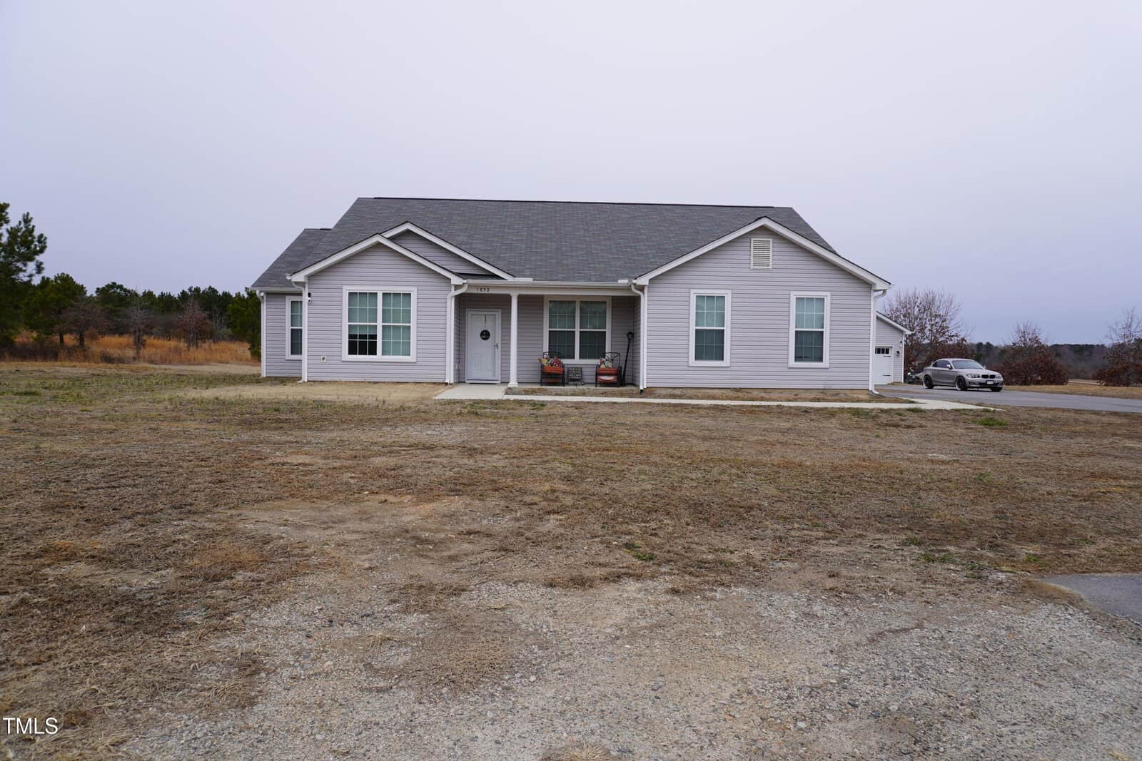 The single-story gray house at 1850 Gillburg Road in Henderson features a dark roof and rests on a sparse, grassy lawn. It boasts white-framed windows, a front door with a wreath, and a driveway to the right with a car parked at the edge. Sparse trees stand under an overcast sky.
