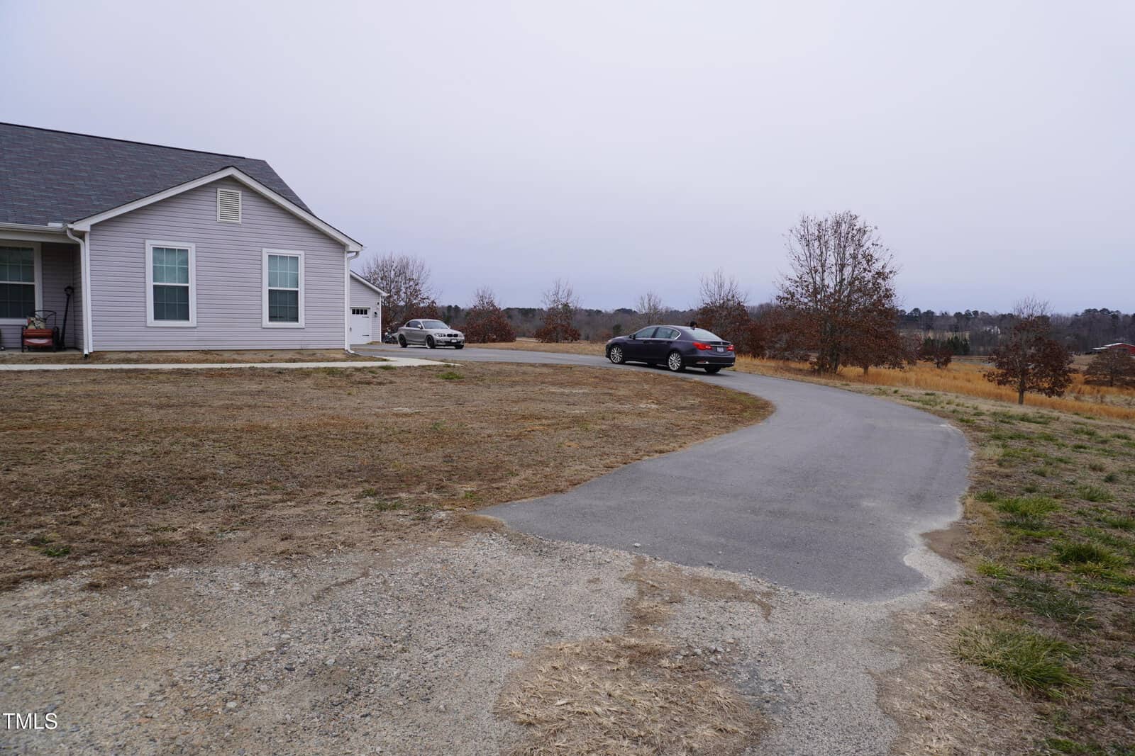 A suburban house with a light-colored exterior is situated at the end of a curved driveway. Several cars are parked near the house. The yard is mostly dry grass with a few small trees. The sky is overcast.