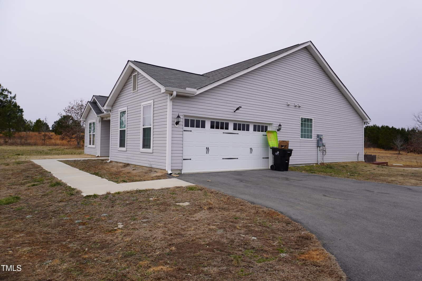 Single-story gray house with a gabled roof, a double garage door, and a paved driveway. A sidewalk leads to the side entrance. The yard is dry and sparse, and a couple of trash bins are near the garage. Overcast sky.