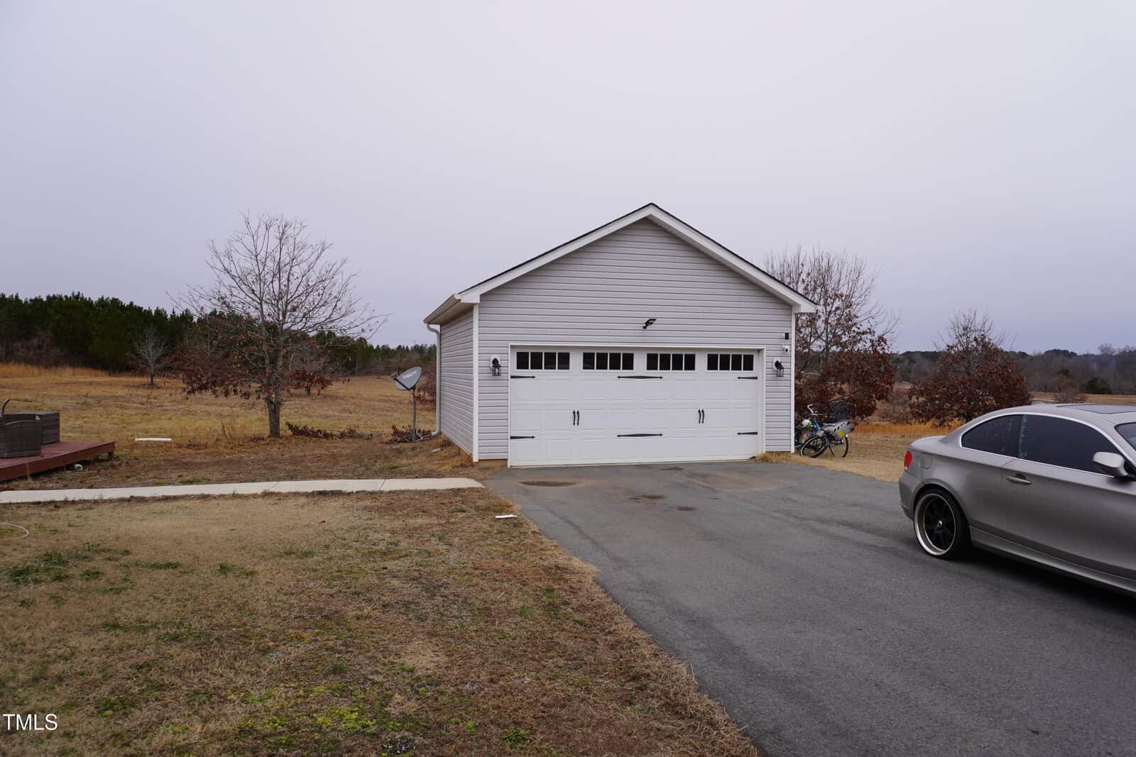 A single-car garage with a white exterior and two closed doors is situated on a paved driveway. A silver car is parked in front. The surrounding landscape features dry grass, leafless trees, and an overcast sky. A bicycle is visible near the garage.