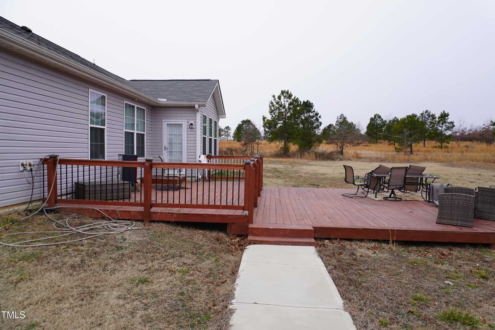 Wooden deck attached to a light-colored house with large windows. It features a dining table and chairs. A hose is coiled on the deck. A concrete pathway leads to the deck, and there is an open grassy area with trees in the background.