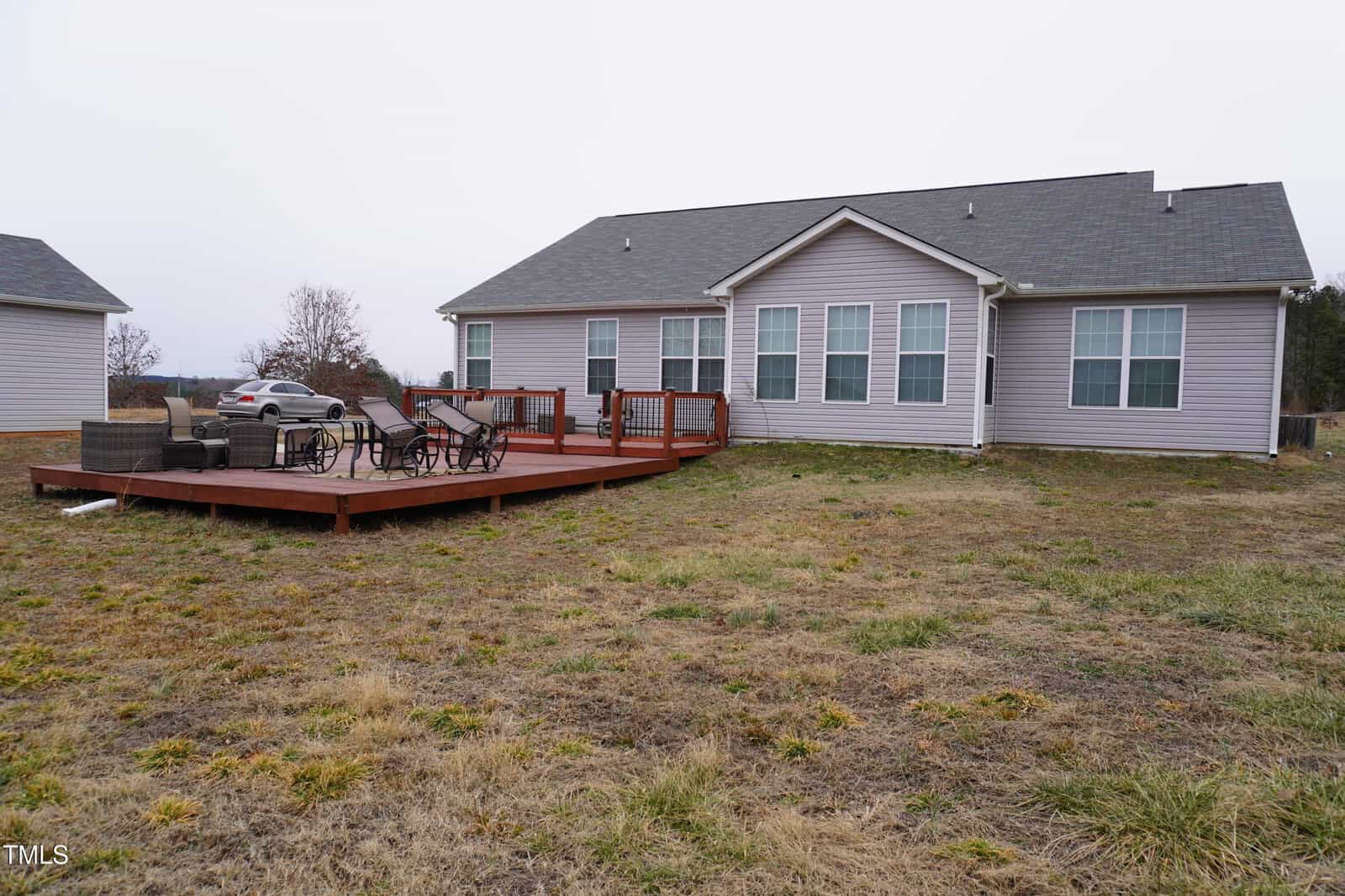 Single-story gray house with white trim and multiple windows is shown from the backyard perspective. A wooden deck features outdoor seating and a grill. The yard is mostly grass with some patches of dirt. The sky is overcast.