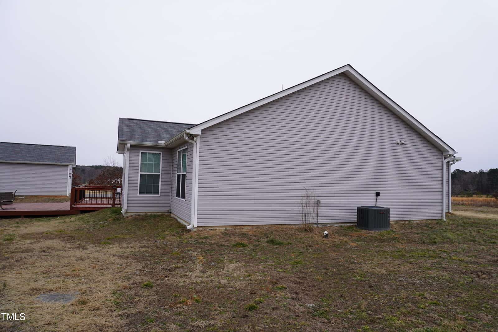 The image shows the side view of a gray, single-story house with a gabled roof and a deck attached to the back. The siding is vinyl, and there is a small air conditioning unit at the rear. The yard is grassy and appears slightly overgrown.