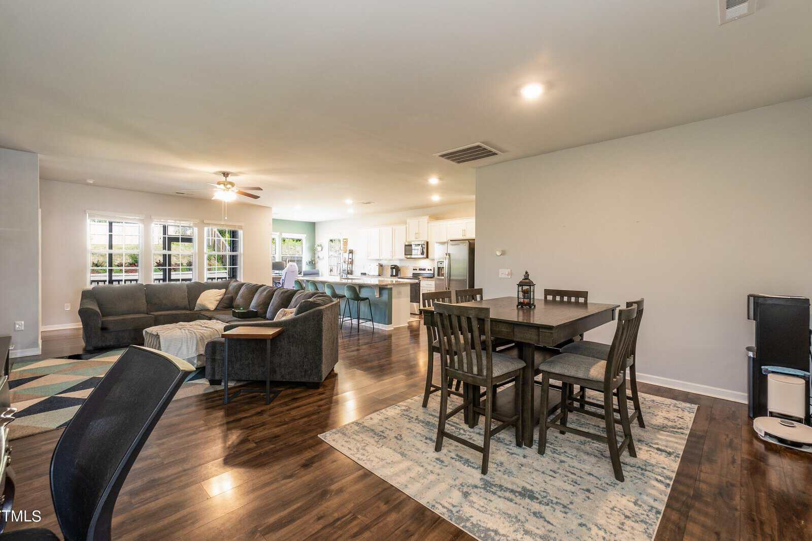 Open-plan living room with dark wood flooring, featuring a large gray sectional sofa, black dining table with chairs, and a kitchen with white cabinets and stainless steel appliances. Large windows in the background offer natural light.