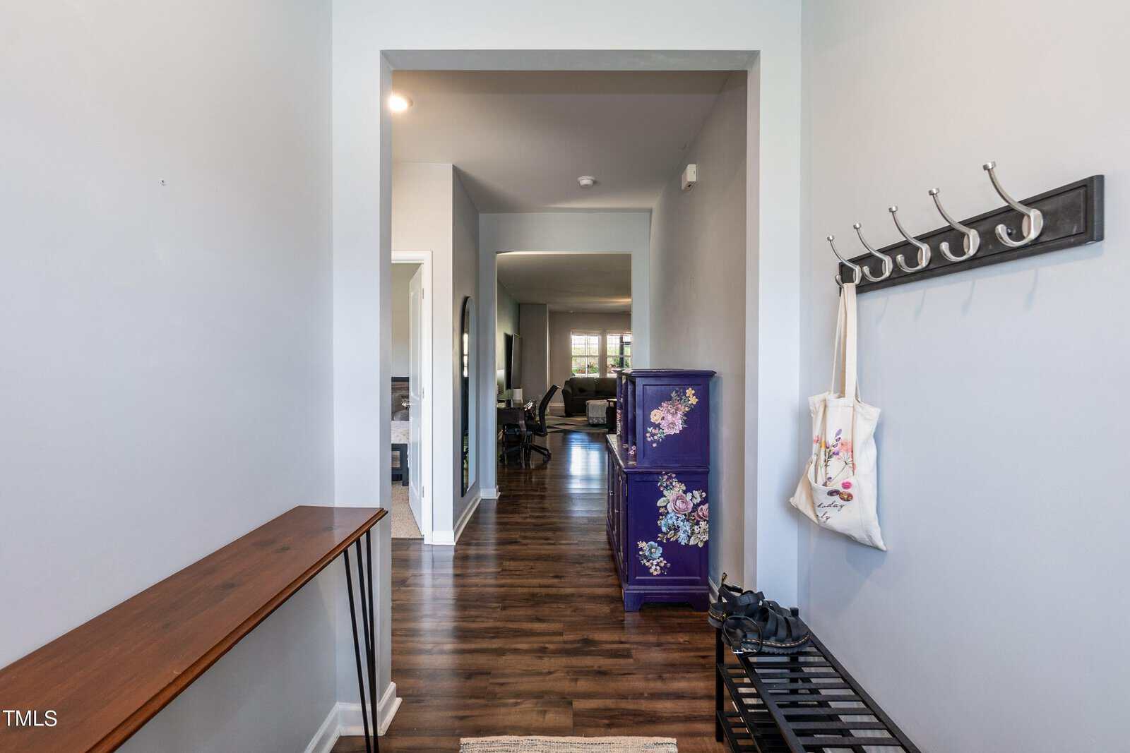 A modern hallway with wooden floors, a coat rack with a bag hanging, and a bench with shoes. A purple cabinet with floral designs stands on the right, leading to a living area visible in the background.