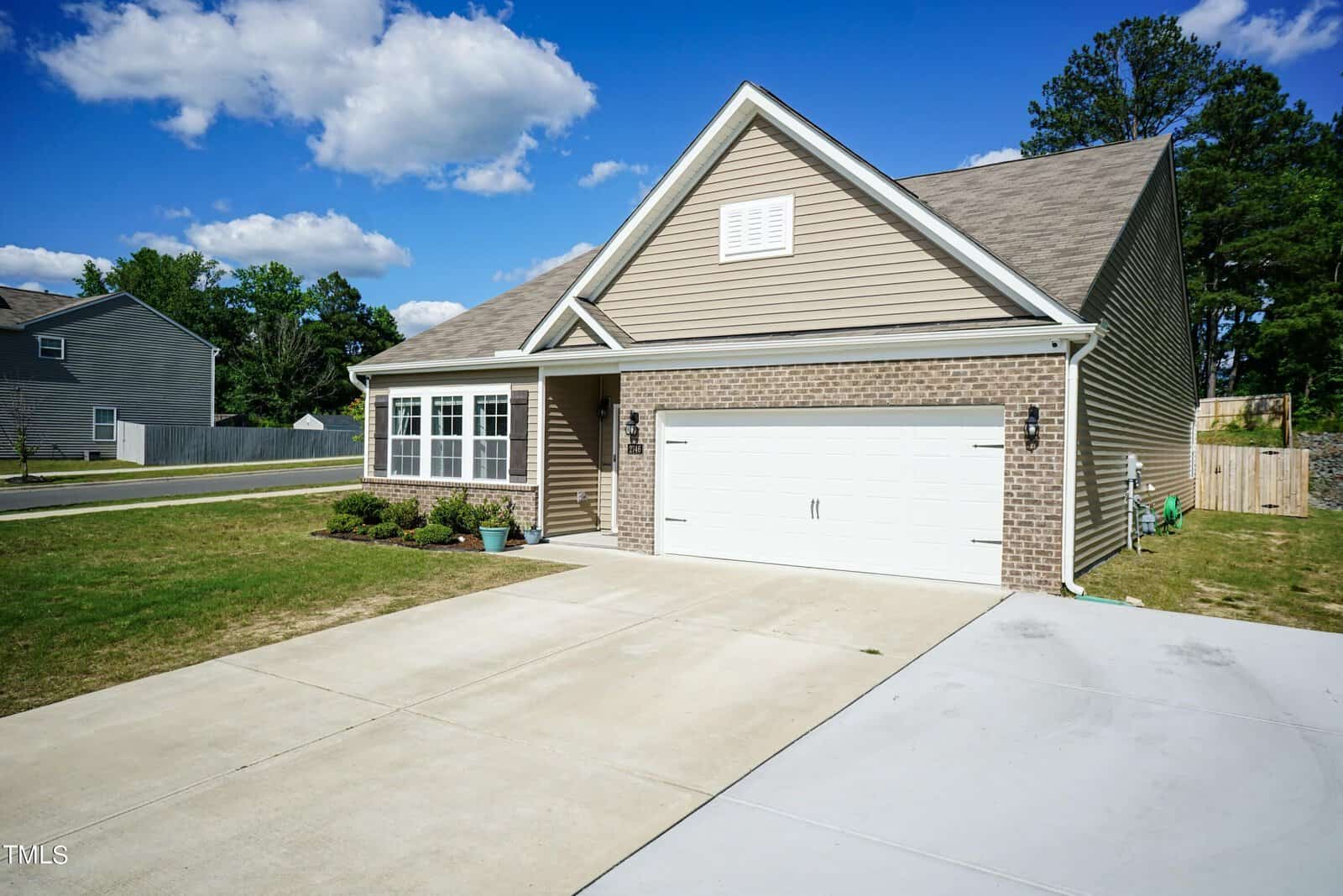 A modern single-story house with a brick facade and beige siding, featuring a two-car garage and a well-maintained front yard. A concrete driveway leads to the garage, and the sky above is partly cloudy.