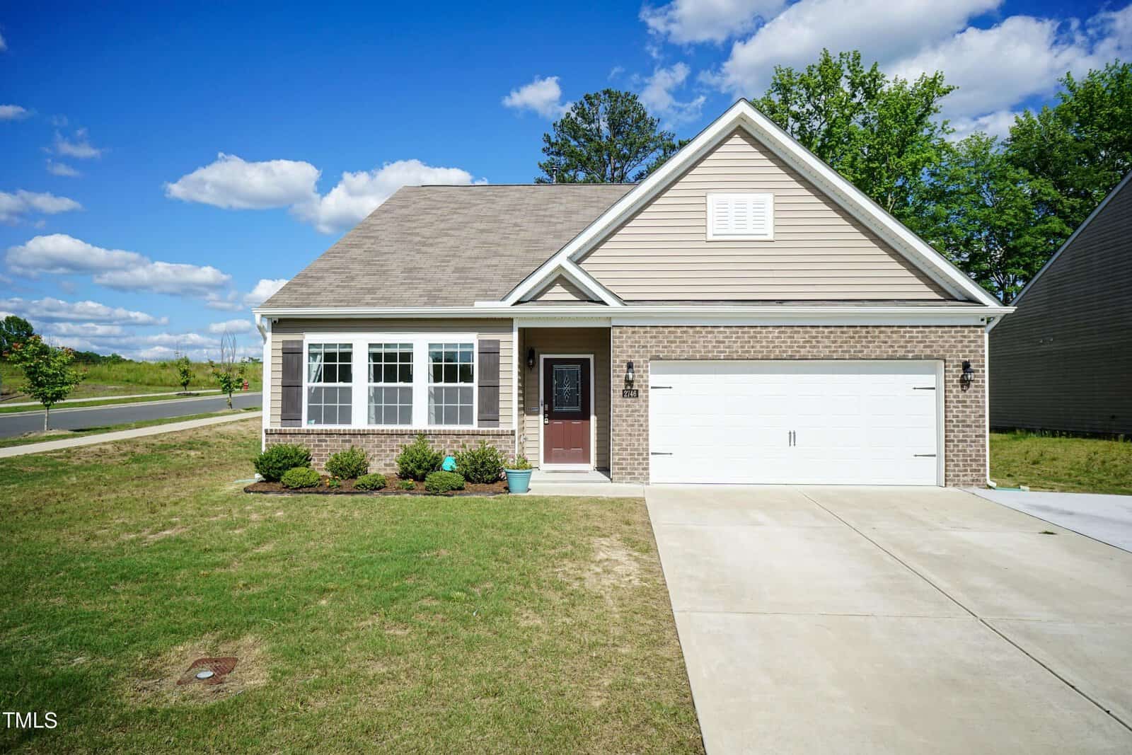 A single-story house with beige siding and a large white garage door. The front yard features a neatly manicured lawn and a small garden. A clear blue sky with scattered clouds serves as the backdrop.