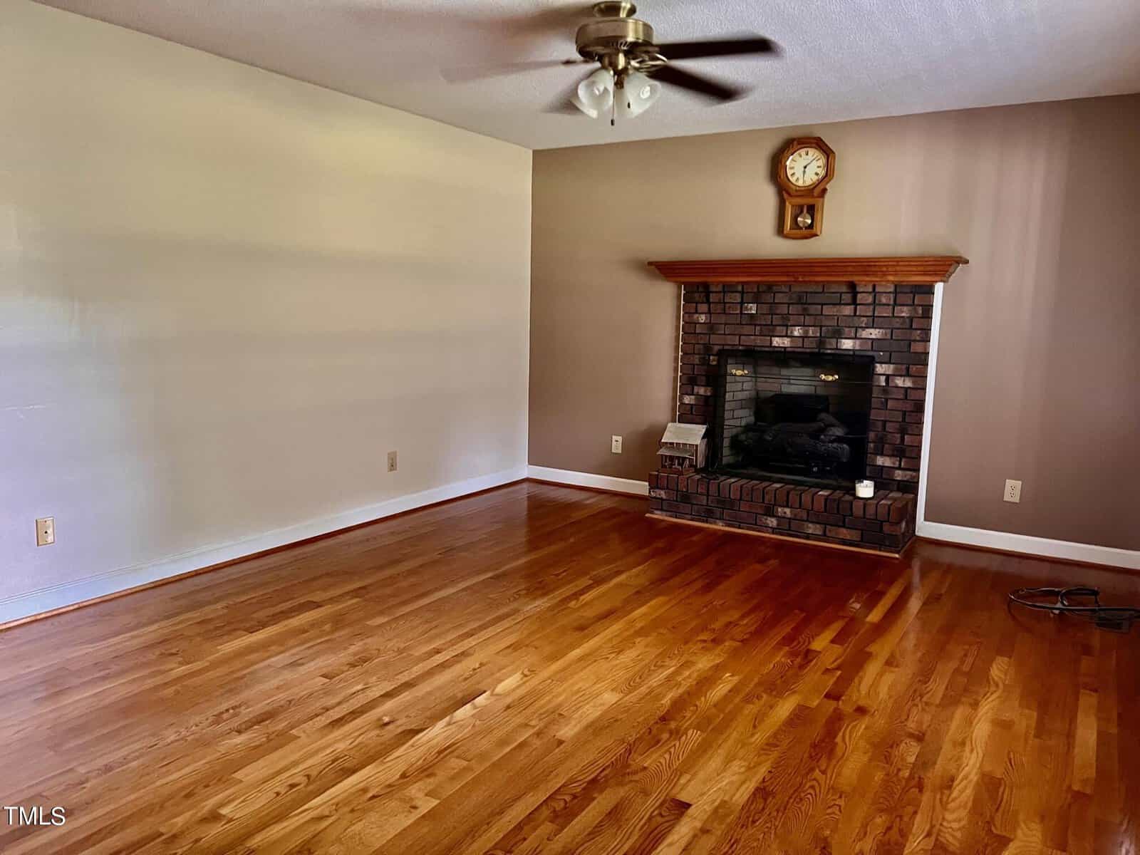 A vacant living room with wooden floors and beige walls. A ceiling fan is above, and a brick fireplace with a wooden mantel is on one wall. A clock and small decorative items are on the mantel. No furniture is present.